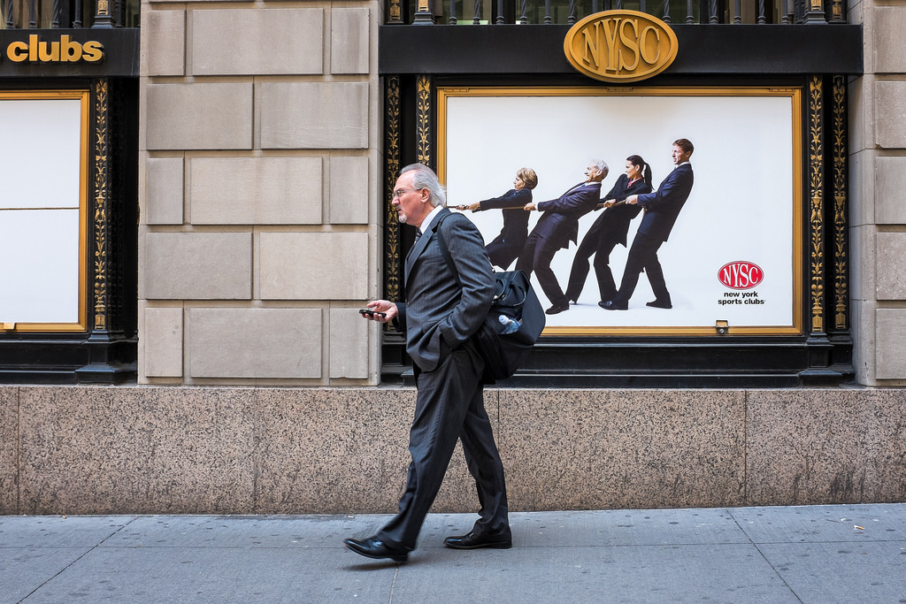 "Wall Street" May 2014 , New York City Fujifilm X100s, 23mm, f/5.6, 1/150, ISO 800 I took this photo on Wall Street, a block east of the New York Stock Exchange. While I'd like to tell you that this was a single, serendipitous moment, it didn't quite come together that way. After noticing the poster on the wall and getting the idea for the photo, I went back to this spot for two days in a row trying to capture this image. It must have taken 200 tries before I got one with a single businessman walking in the right direction, looking just the right amount of unhappy, who was just the right height so the rope would intersect the back of his neck , and frozen at just the right moment so those two inches of rope would actually attach to his collar. 