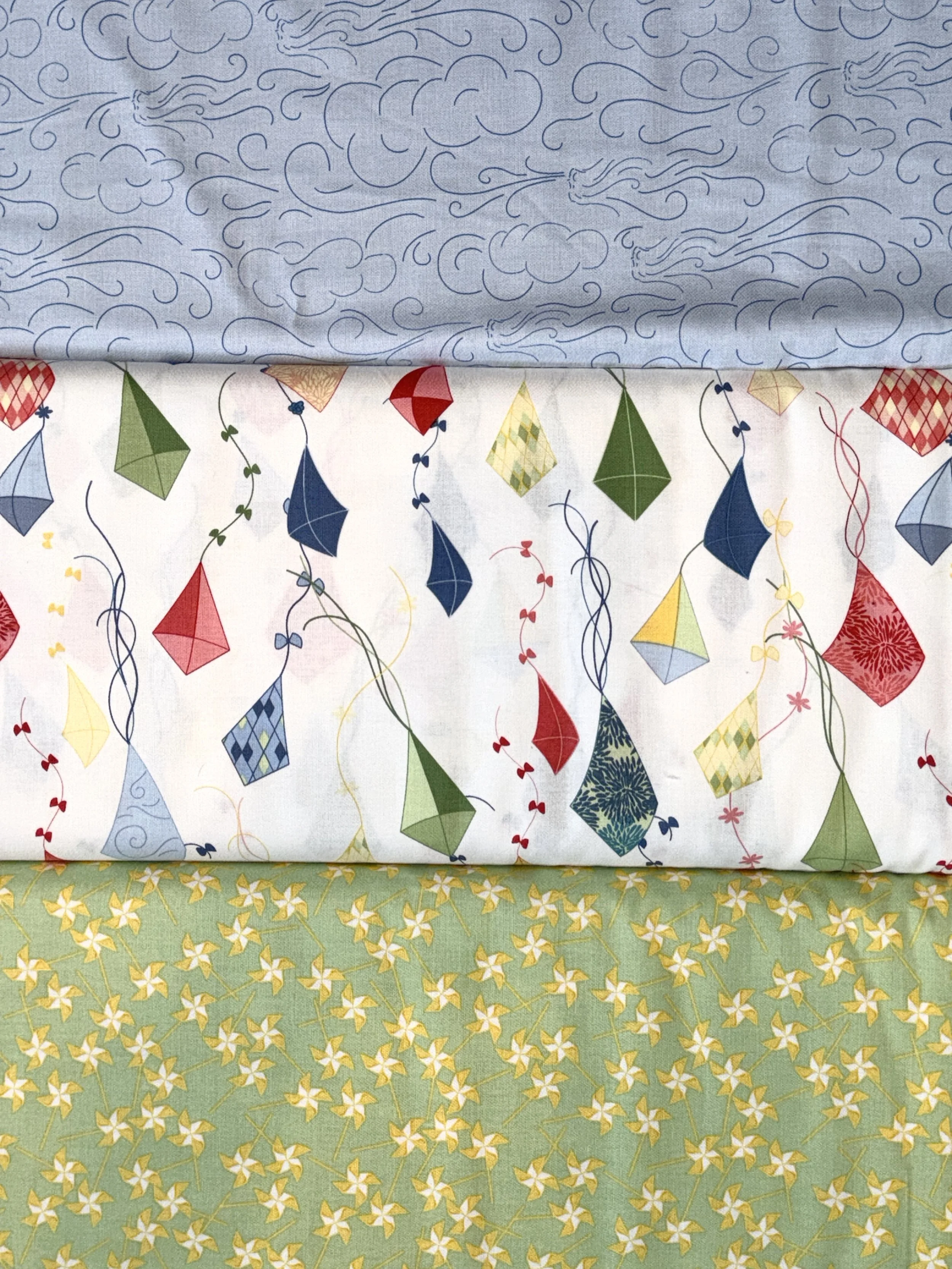 Three coordinating 100% cotton quilting fabrics featuring blue wind clouds, colorful kites on white, and small yellow flowers on a soft green background.