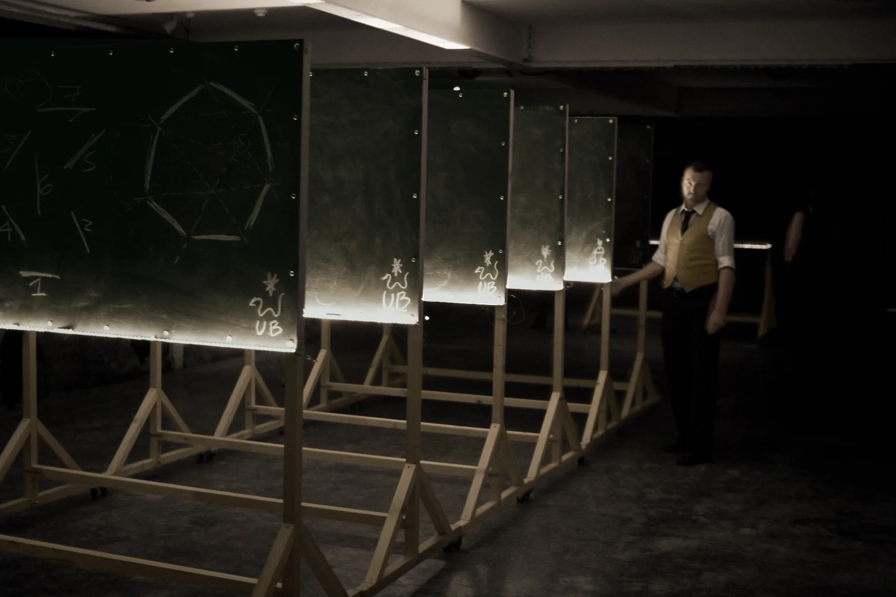 Ulysses Black, as Lecturer 1, reveals the presence of the linear sequence of blackboards within the first configuration. Performance at the Basement, Brighton 2013