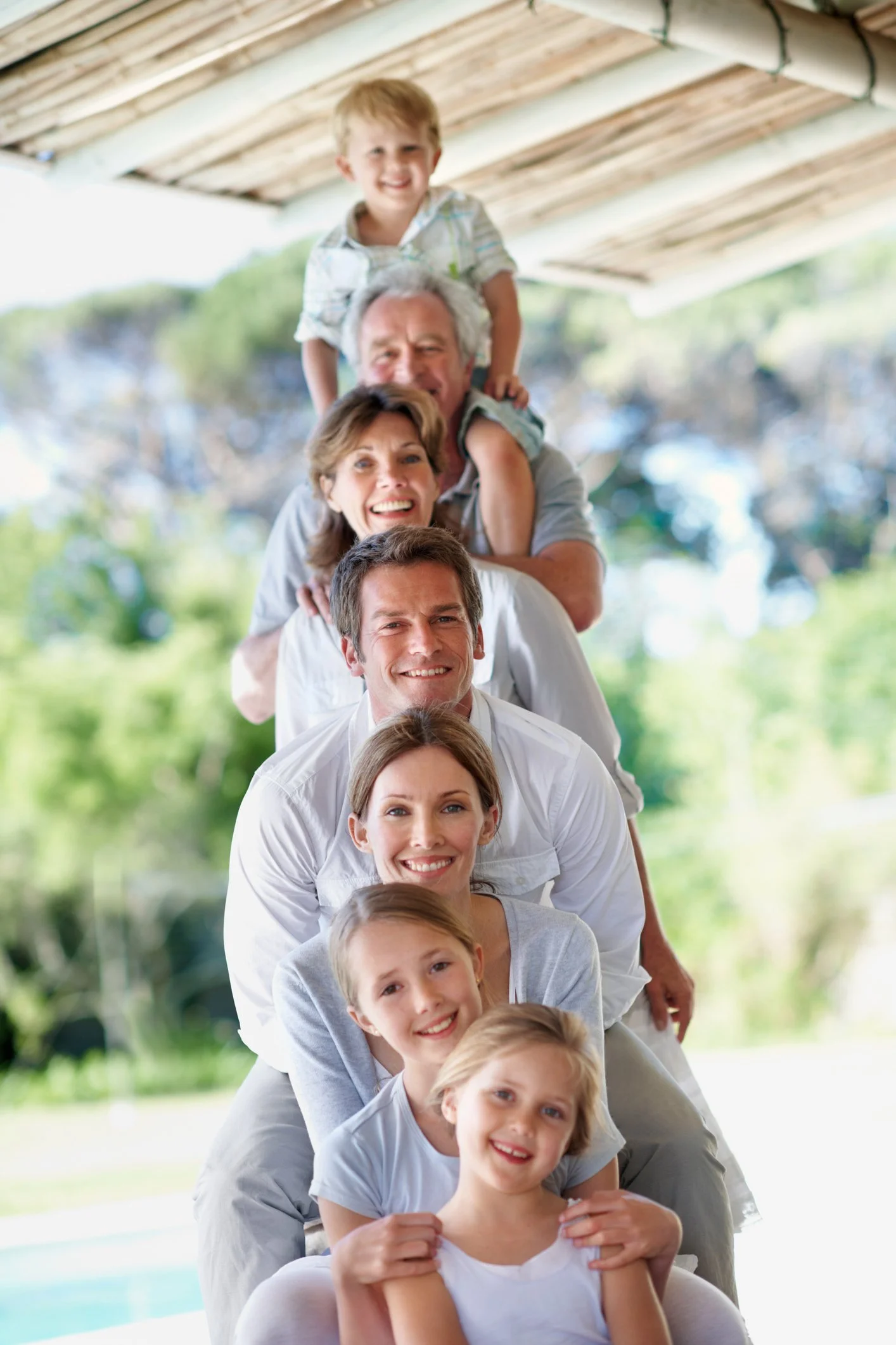 A multigenerational family stacked in a human pyramid outdoors, smiling and enjoying a sunny day with blurred trees and a pool in the background.