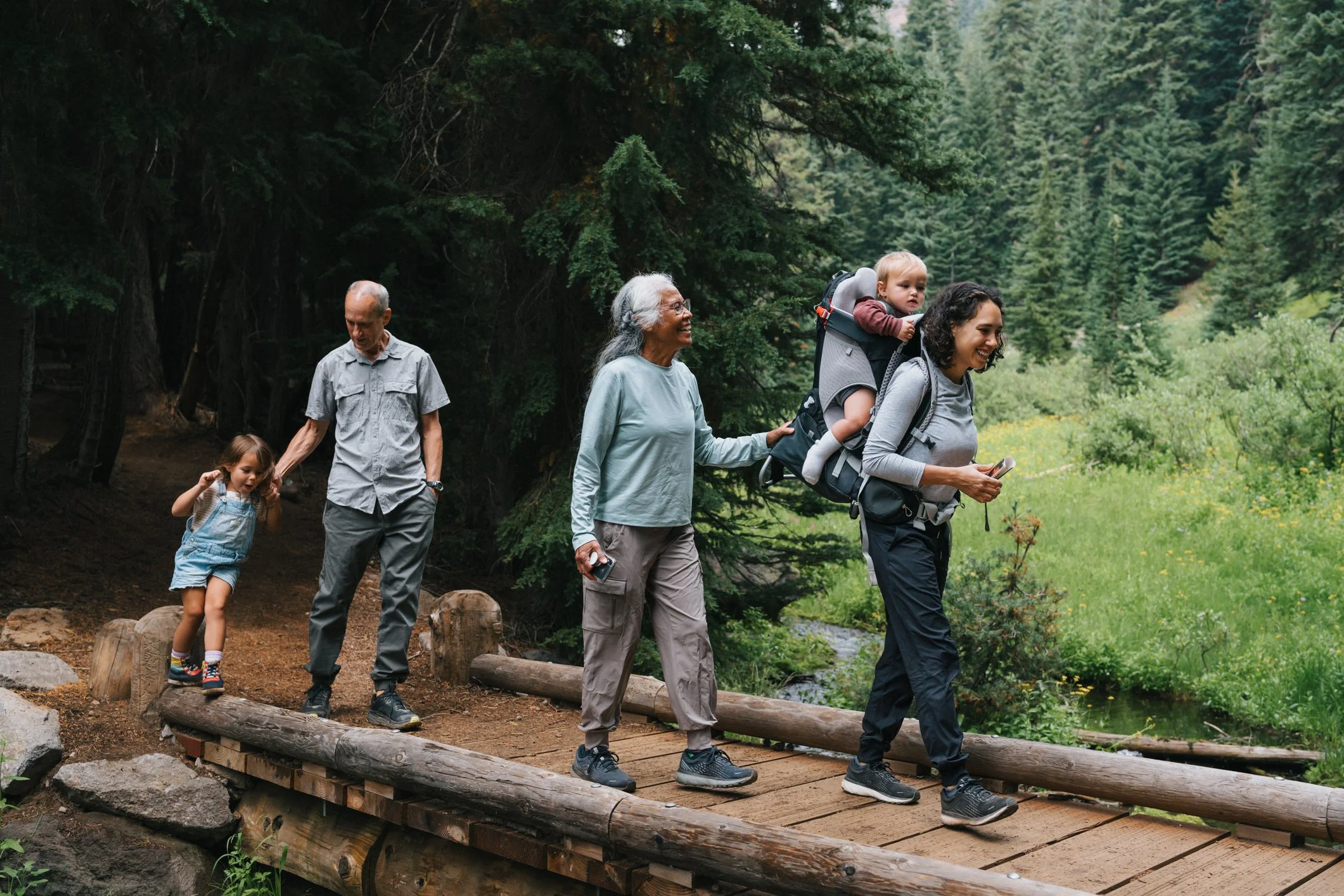 A family of five hiking on a trail in a lush, green forest. The group includes an elderly woman, an elderly man, a young woman with curly hair carrying a small child in a backpack carrier, and a young girl. They are walking across a wooden bridge over a small creek, surrounded by tall trees and dense foliage.