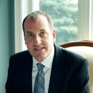 A man in a dark suit and tie sitting indoors near a window, with green trees visible outside.