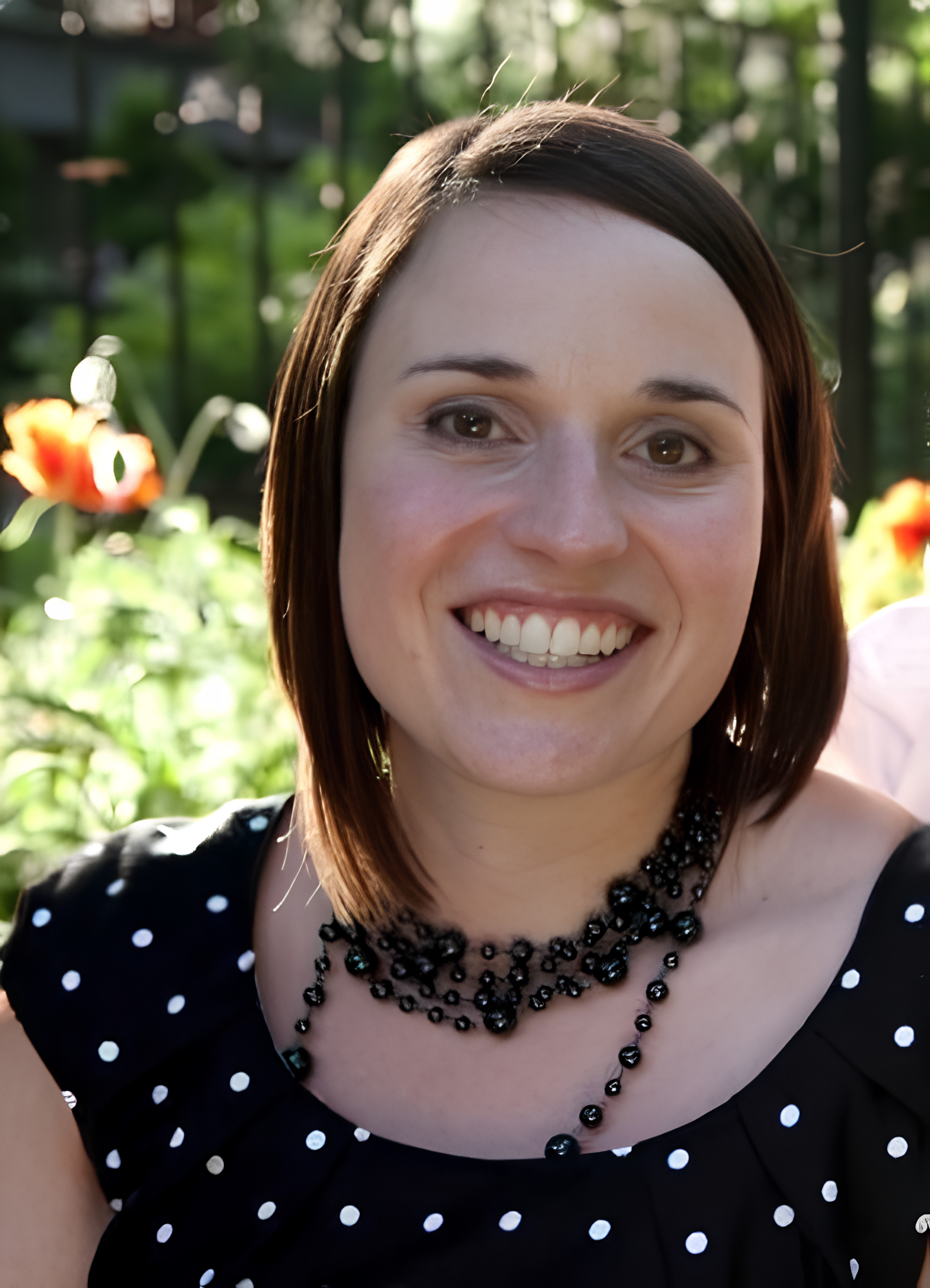 Close-up of a smiling woman with short brown hair, wearing a black dress with white polka dots and a black beaded necklace, outdoors with greenery and flowers in the background.