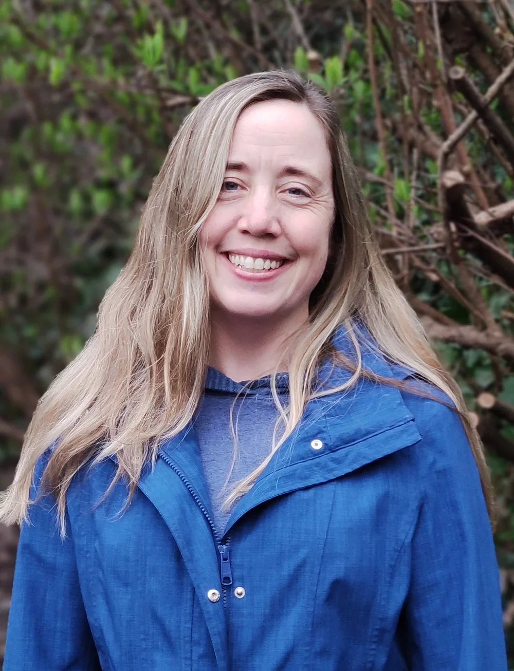 A smiling woman with long, wavy blonde hair wearing a blue jacket outdoors with green foliage and branches in the background.