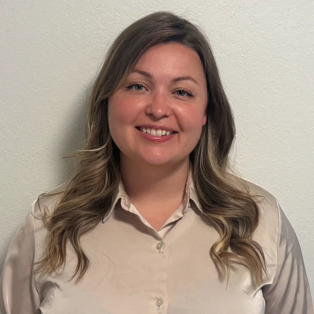 A woman with wavy light brown hair wearing a beige button-up shirt standing against a light-colored wall.