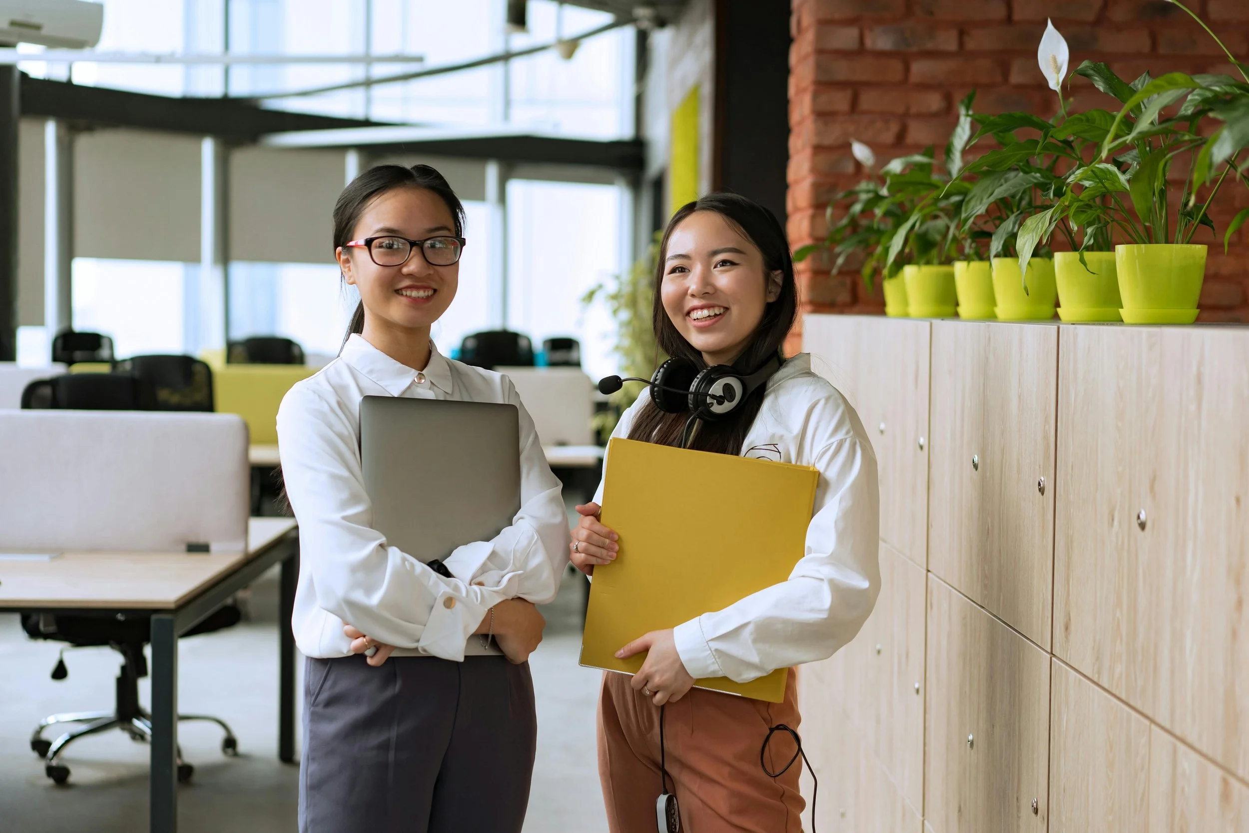 Two women standing together in office space, holding a yellow folder and a laptop, smiling, with potted plants and office furniture in the background.