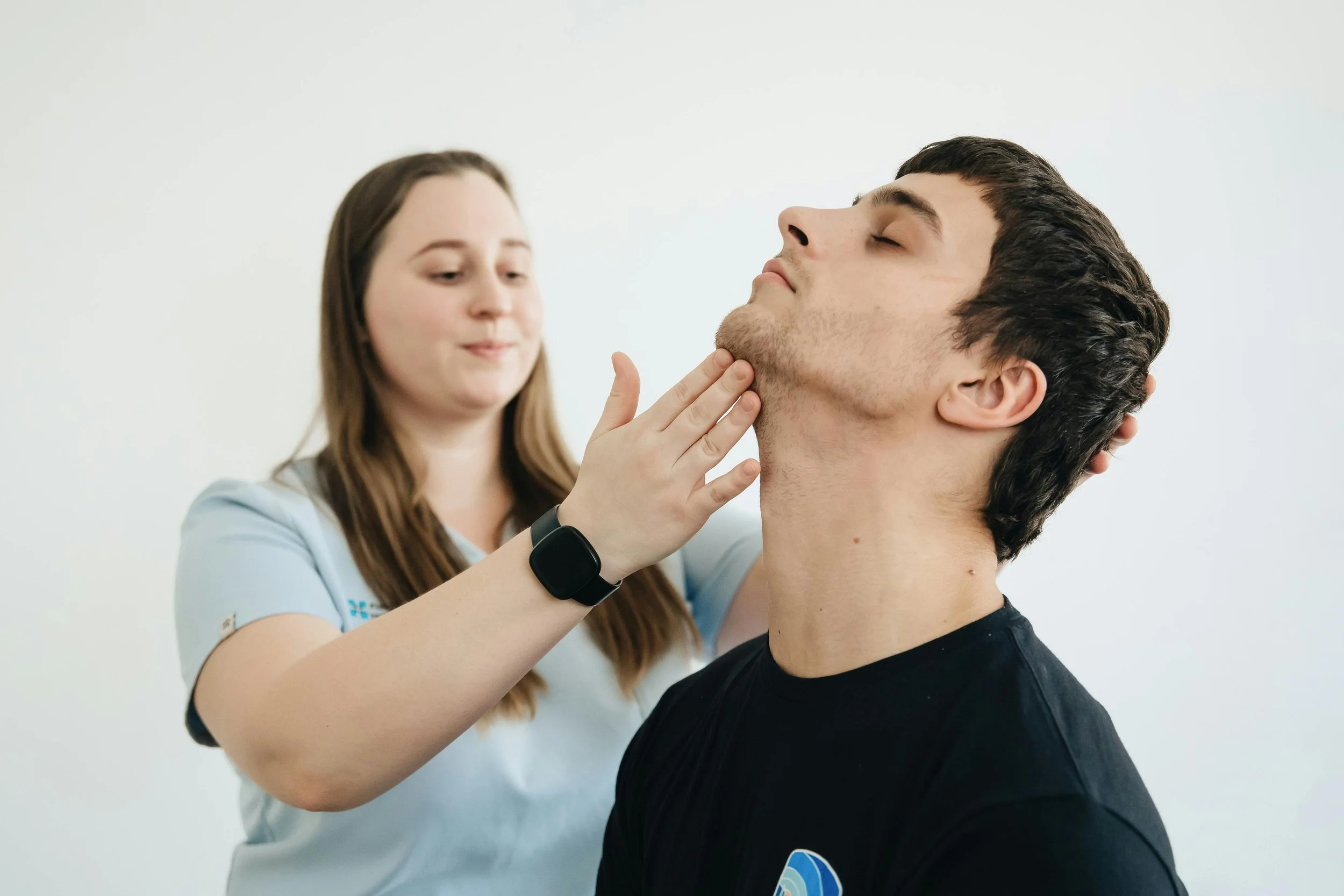 A healthcare professional examines a young man's throat while the young man sits with his eyes closed.