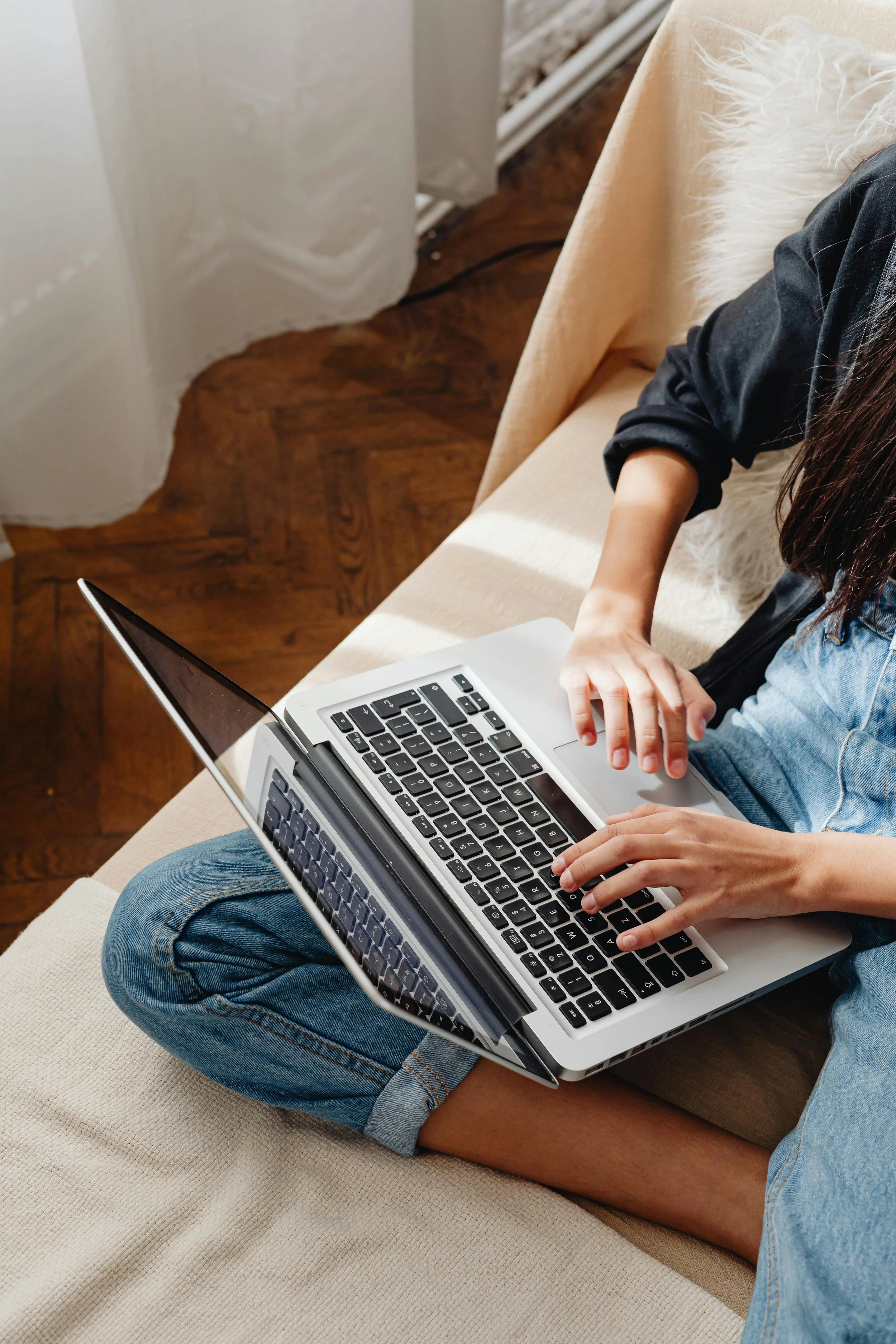 A person sitting cross-legged on a beige couch, using a silver laptop with a black keyboard. They are wearing blue jeans and a dark long-sleeve shirt. Part of the person's head and right arm are visible. The environment includes a wooden floor and a white fluffy throw pillow on the couch.