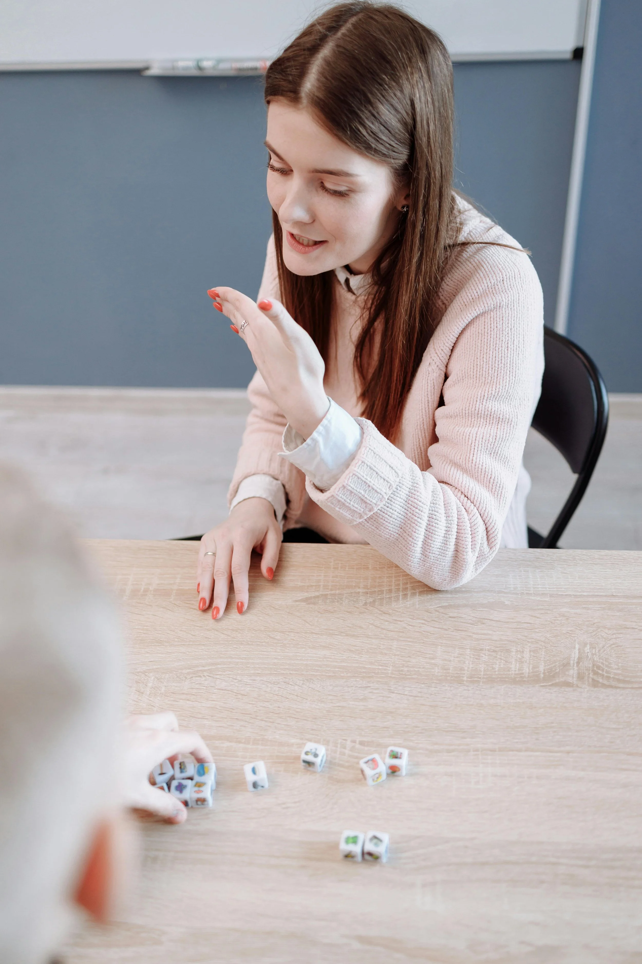 A young woman with long brown hair and a light pink sweater is sitting at a wooden table, engaging in a game of dice with another person. There are several small dice with colorful images on them scattered on the table.