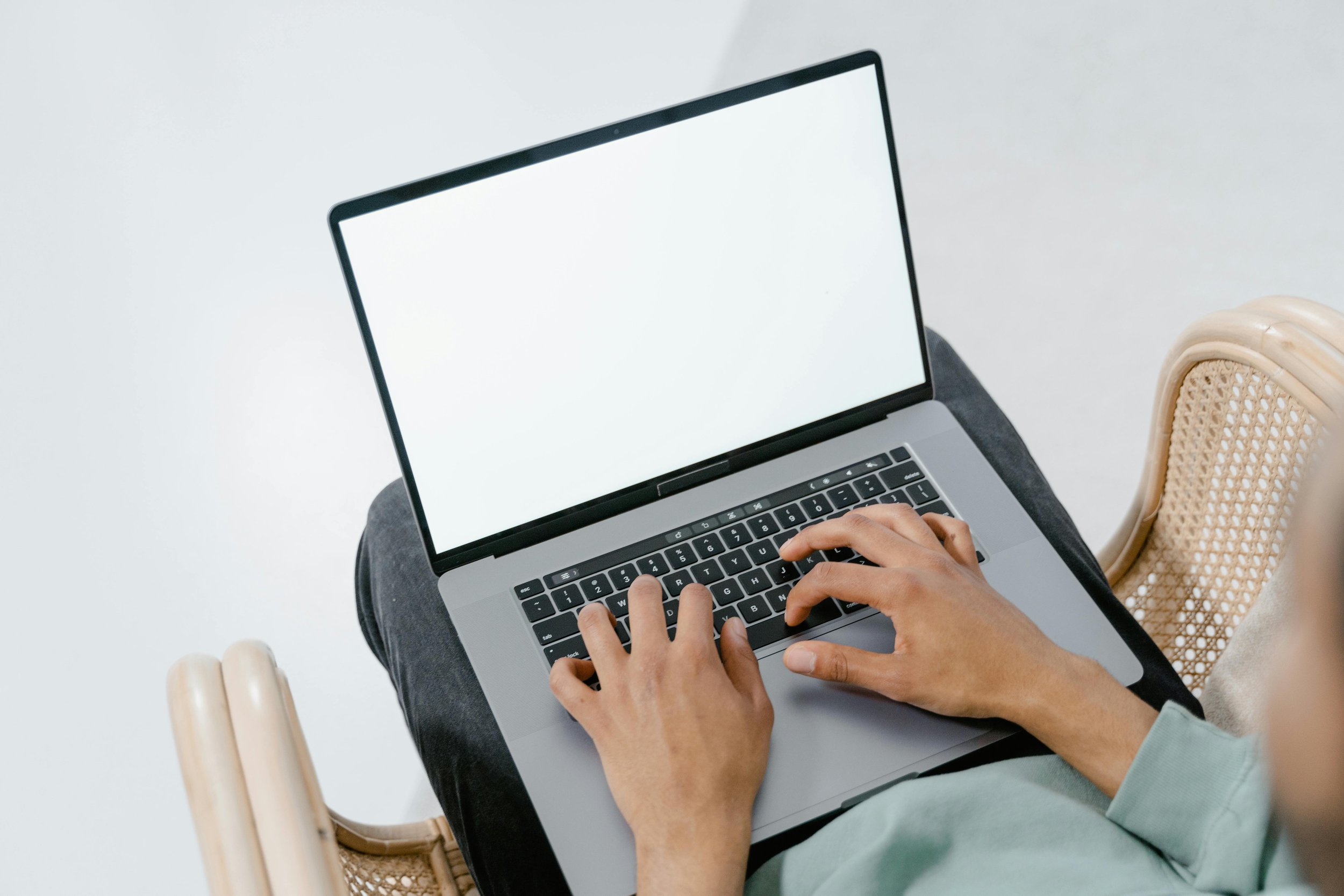 A person typing on a laptop with a blank white screen, sitting in a chair near a wooden armrest.