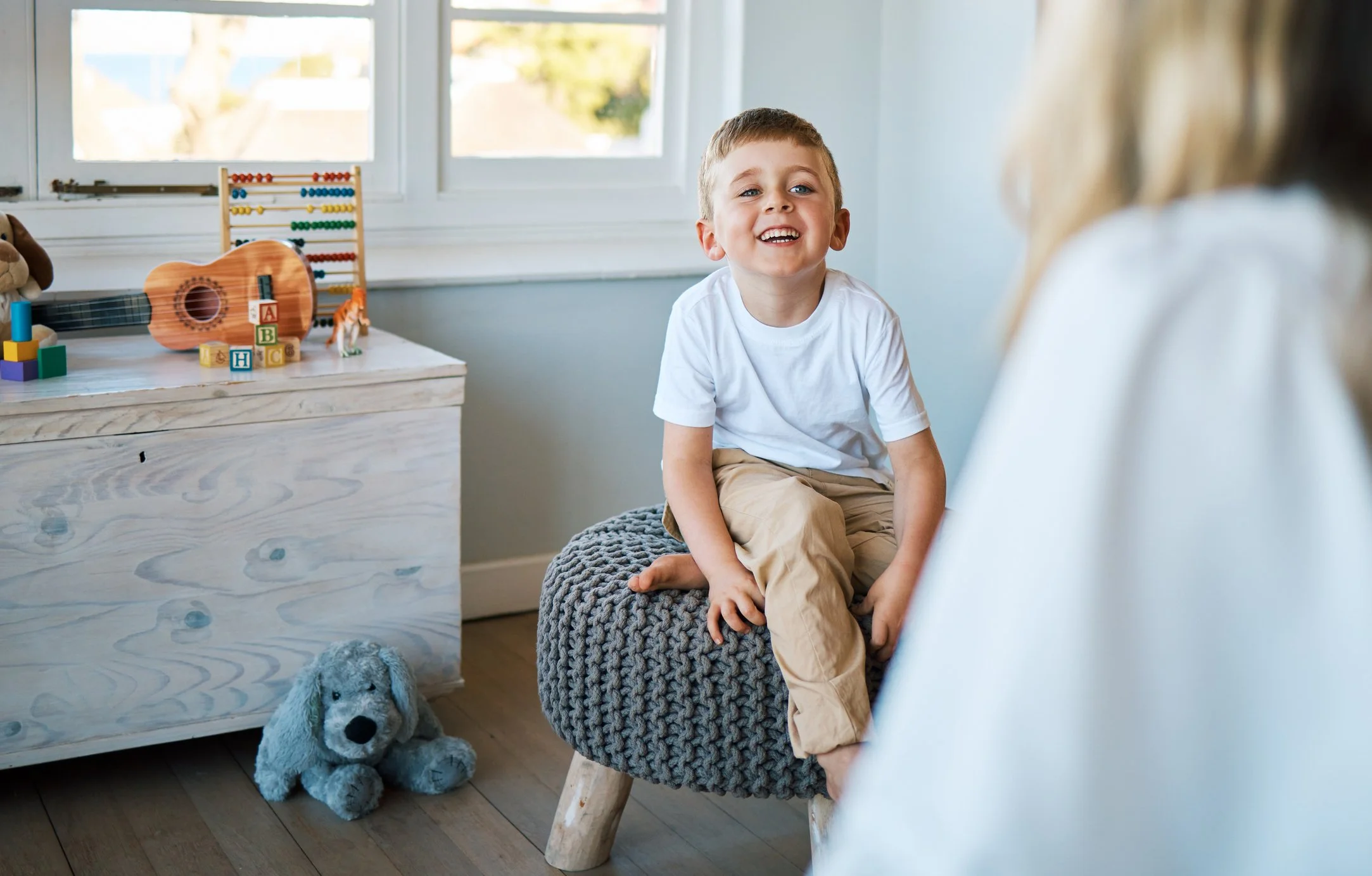 A young boy sitting on a gray knitted stool in a bright room with large windows, smiling and looking at a person in front of him. The room has a white dresser with children's toys including a stuffed dog, a wooden guitar, a colorful abacus, blocks, and a small toy tiger. Near the dresser, there is a plush stuffed dog on the hardwood floor.