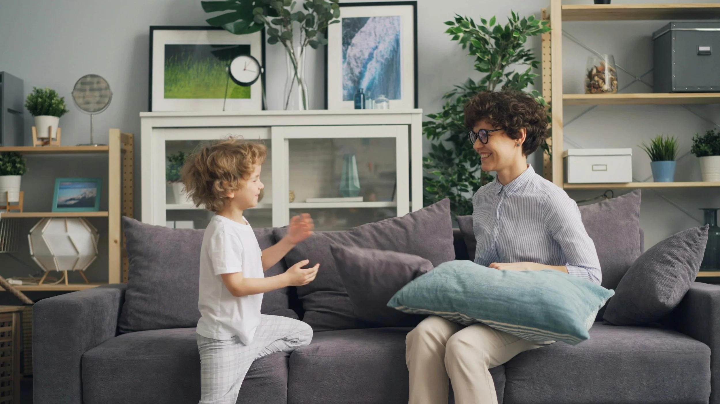 A woman and young girl sitting on a gray couch in a living room, smiling and talking to each other. The woman is wearing glasses and a striped shirt, and the girl is wearing a white shirt and plaid pants.