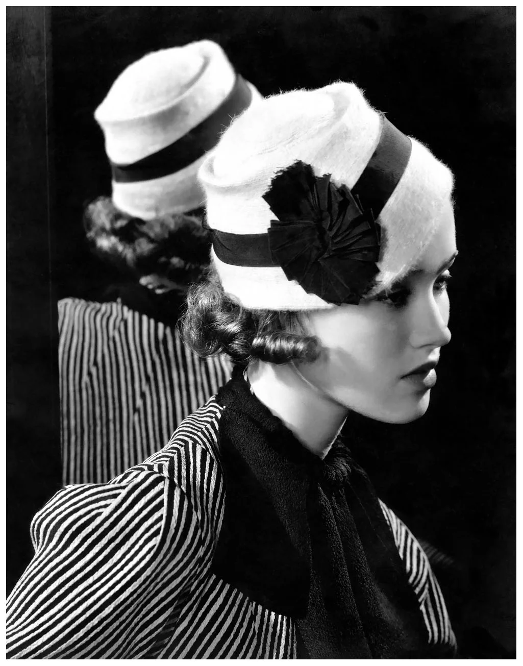 Mary Taylor wearing white angora hat with grosgrain band by Maria Guy, striped rough silk ensemble, and dark shirred scarf, with reflection in mirror behind her 1933