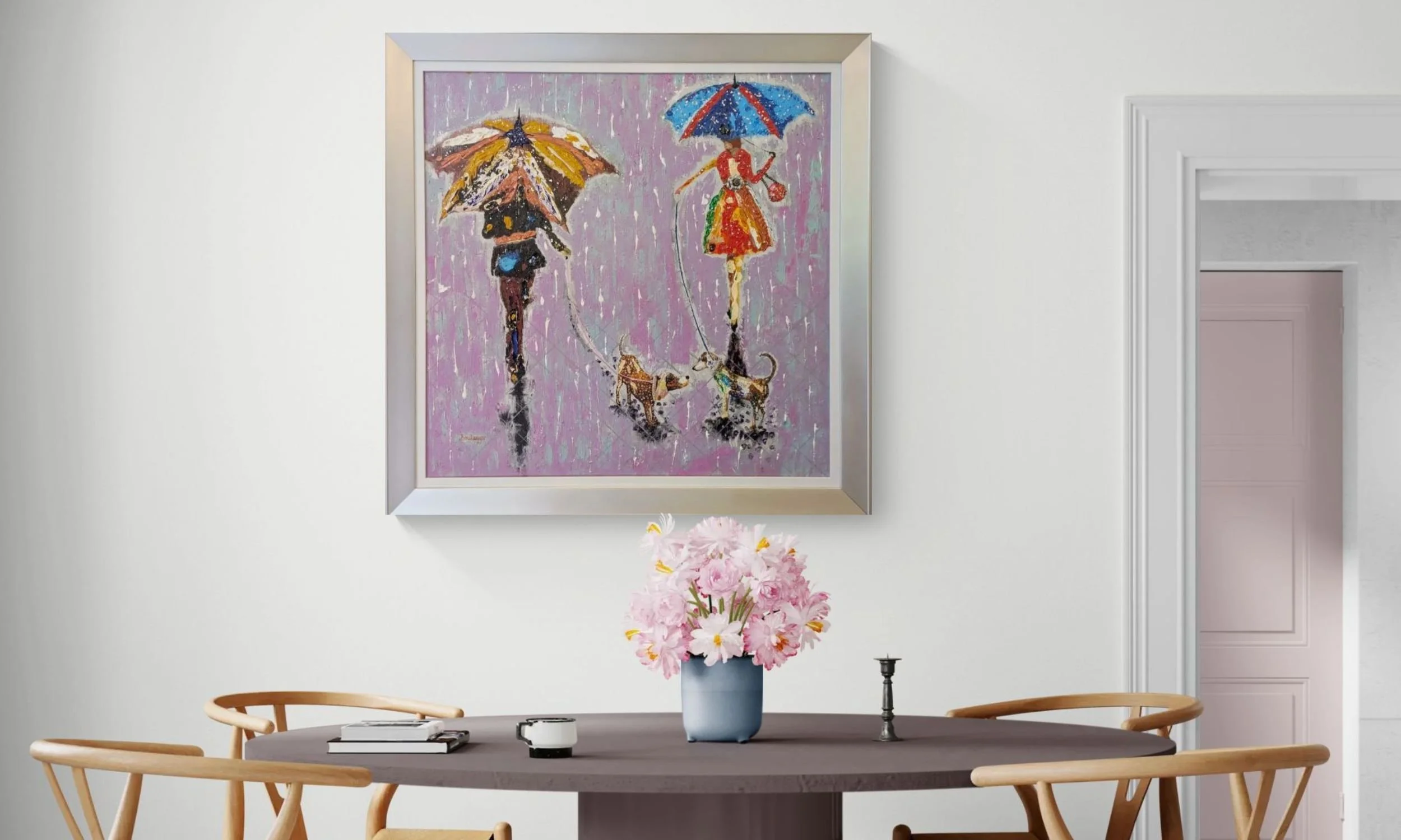 A framed contemporary painting hanging on a light wall, depicting two figures walking two dogs in the rain under umbrellas, placed above a table with a bouquet of flowers in a blue vase.