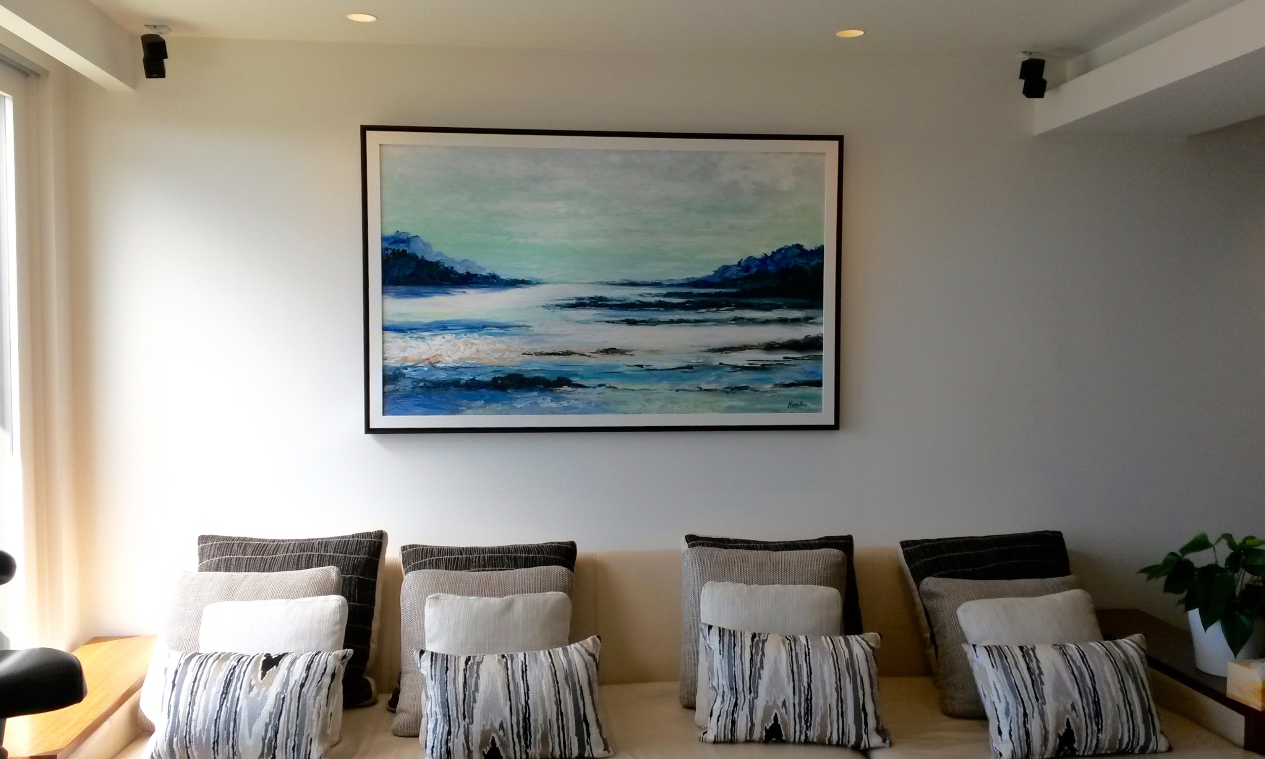 A large horizontal abstract coastal landscape painting with blue and white tones, professionally custom framed in a thin black gallery frame, hanging above a neutral lounge in a Mosman home.