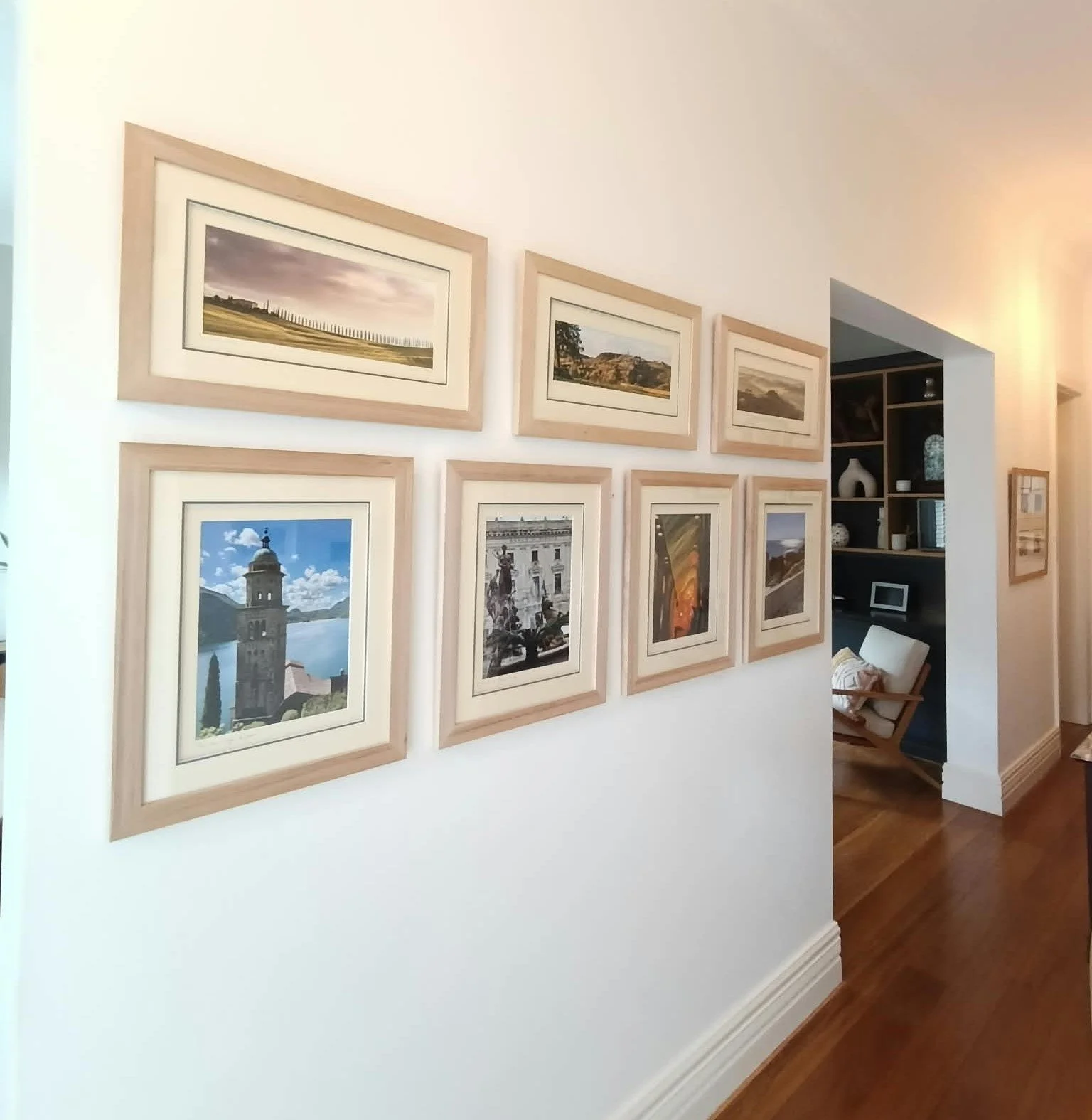 A beautifully arranged hallway gallery wall displaying a collection of landscape and architectural photographs in custom natural timber frames, showcasing professional photo printing and framing services.