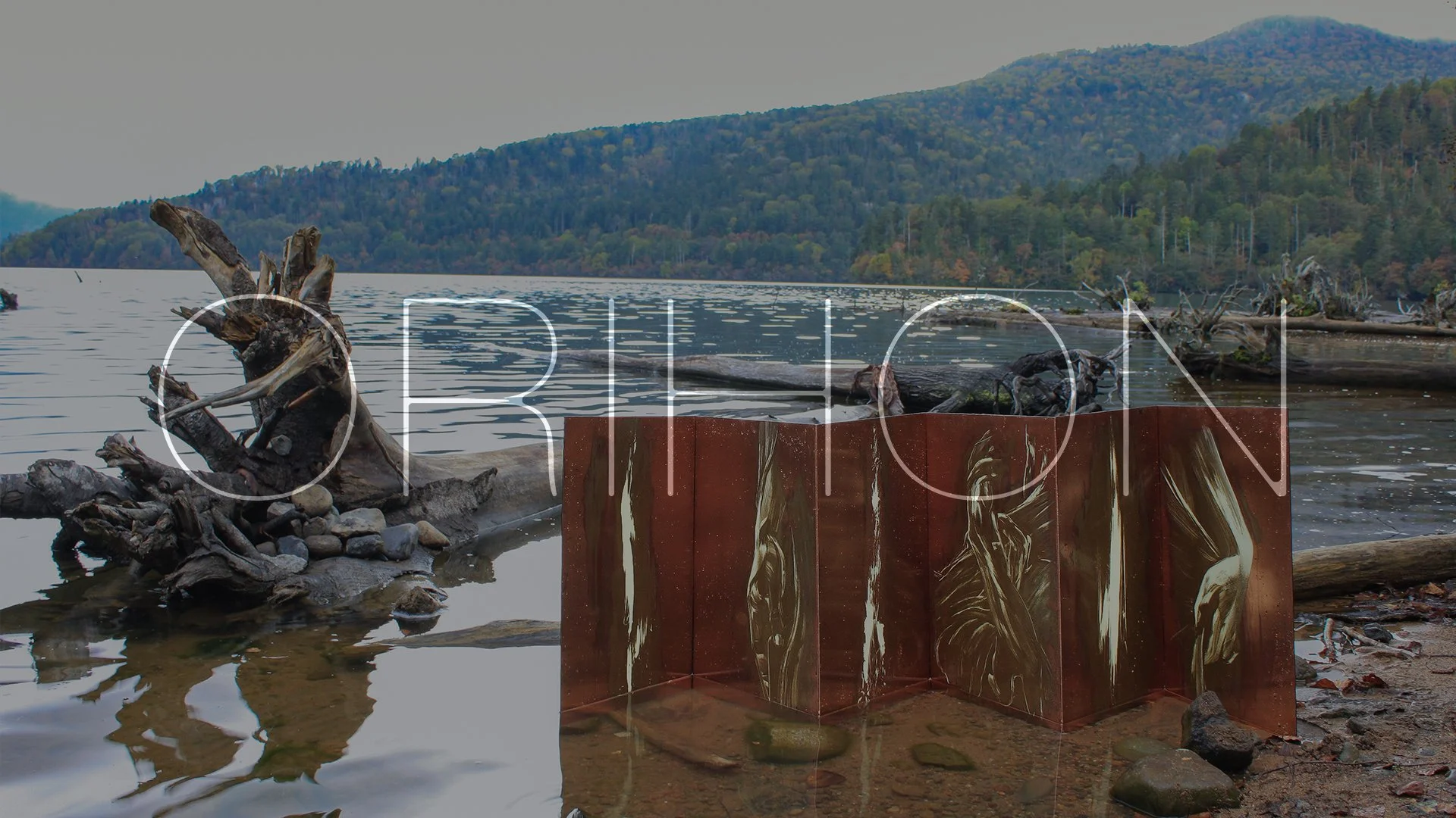 A lakeside scene with a large piece of rust-colored art with metallic swirls in the foreground. There is a driftwood sculpture on the shoreline and a mountainous landscape in the background.