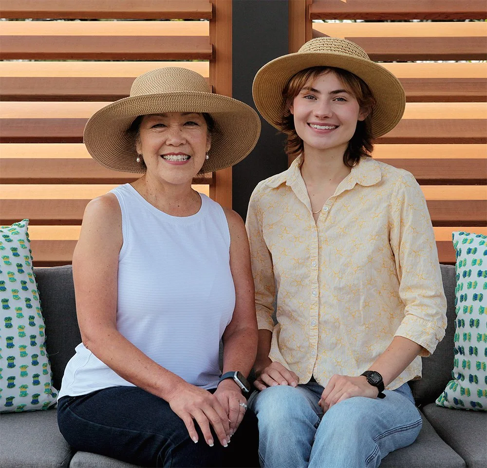 Two people smiling on a couch outdoors, wearing straw hats and light summer clothes.