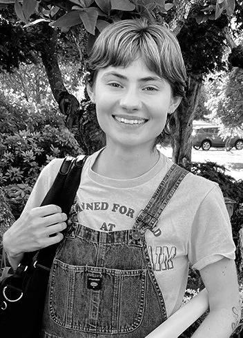 Black and white portrait of a young woman with short dark hair, wearing a white ribbed sweater, outdoors in a parking lot.