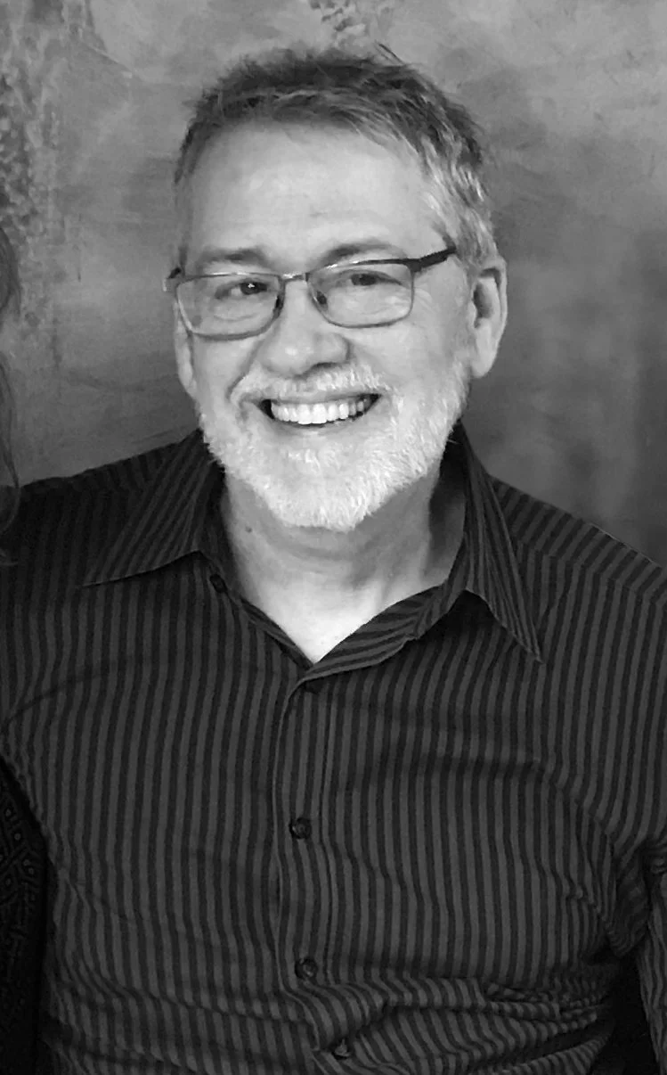 Black and white photo of a smiling man with glasses, gray hair, and a beard.