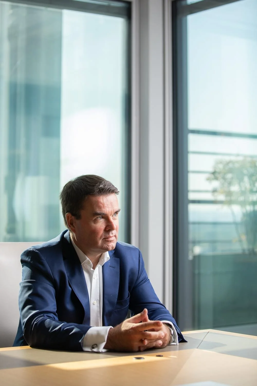 Portrait en situation d'un dirigeant en costume bleu, assis à une table de réunion devant de grandes baies vitrées.