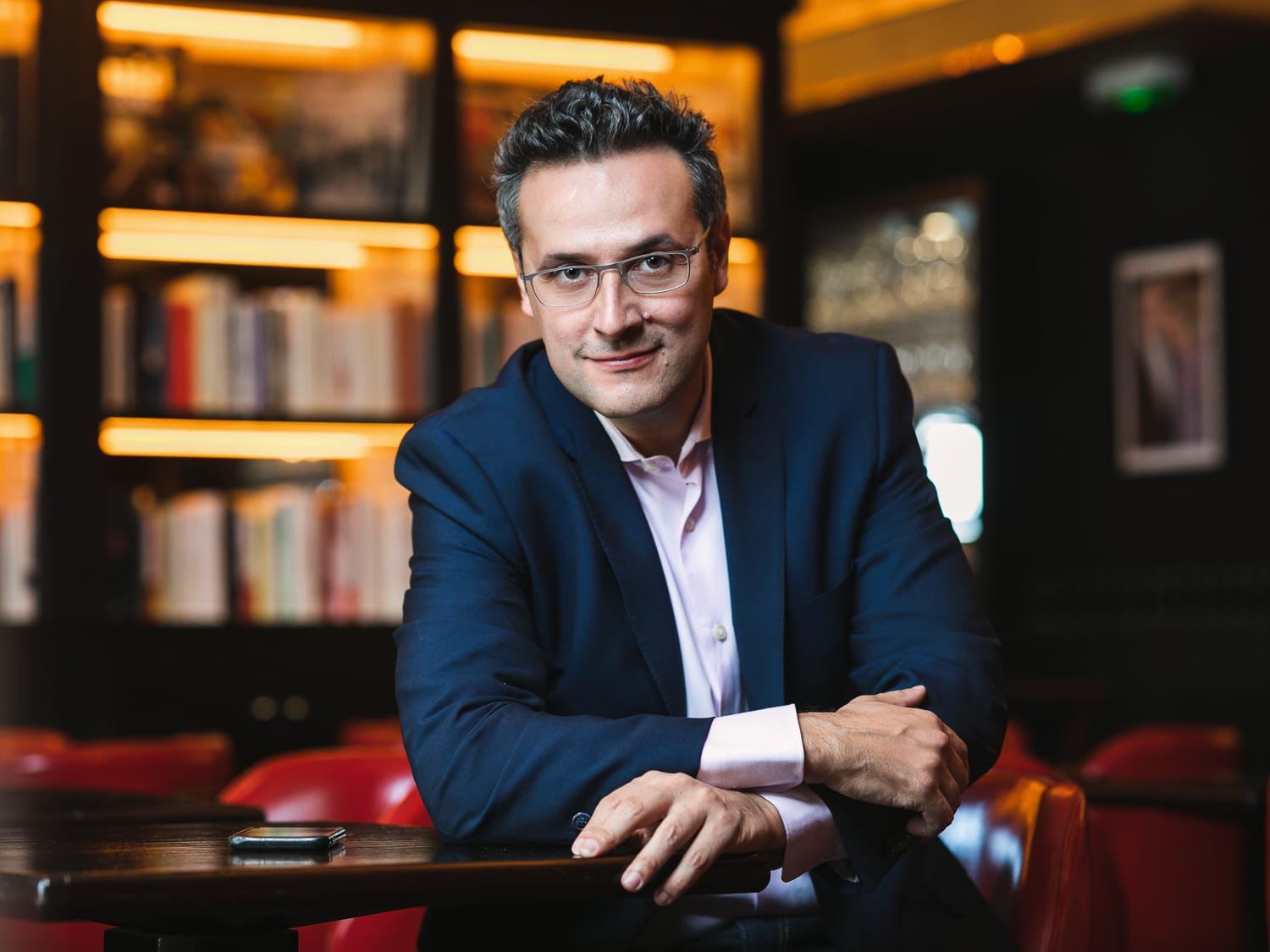 Portrait corporate d'un homme en costume bleu et lunettes, assis devant une bibliothèque aux éclairages chaleureux.