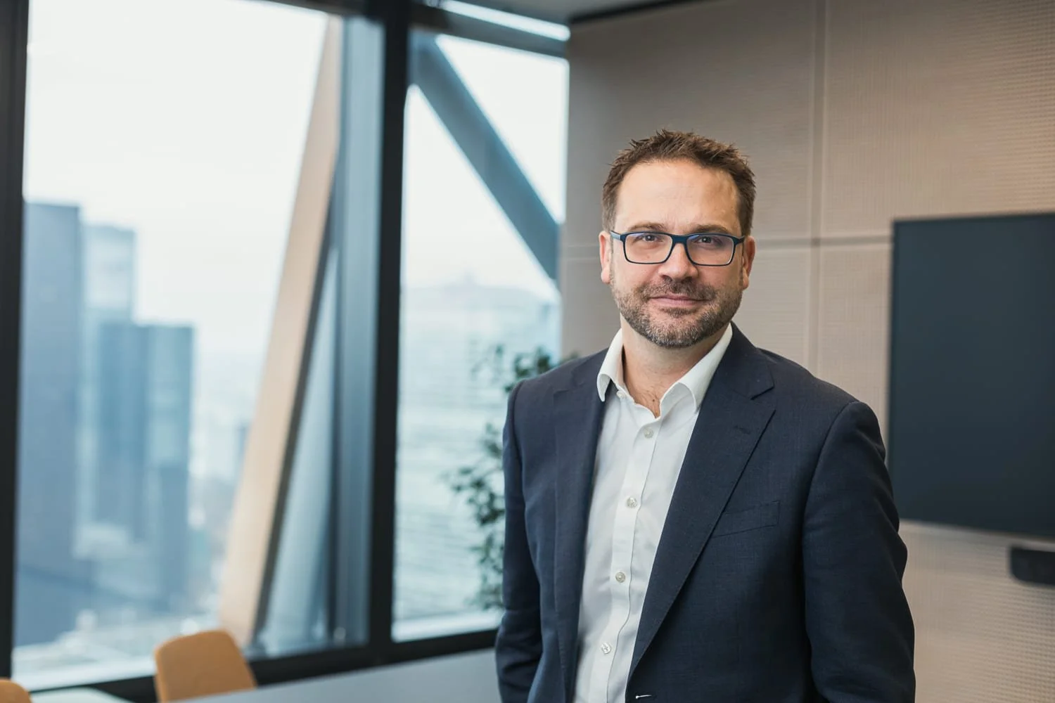 Portrait professionnel d'un homme en costume et lunettes, debout dans des bureaux avec une vue sur les immeubles environnants.