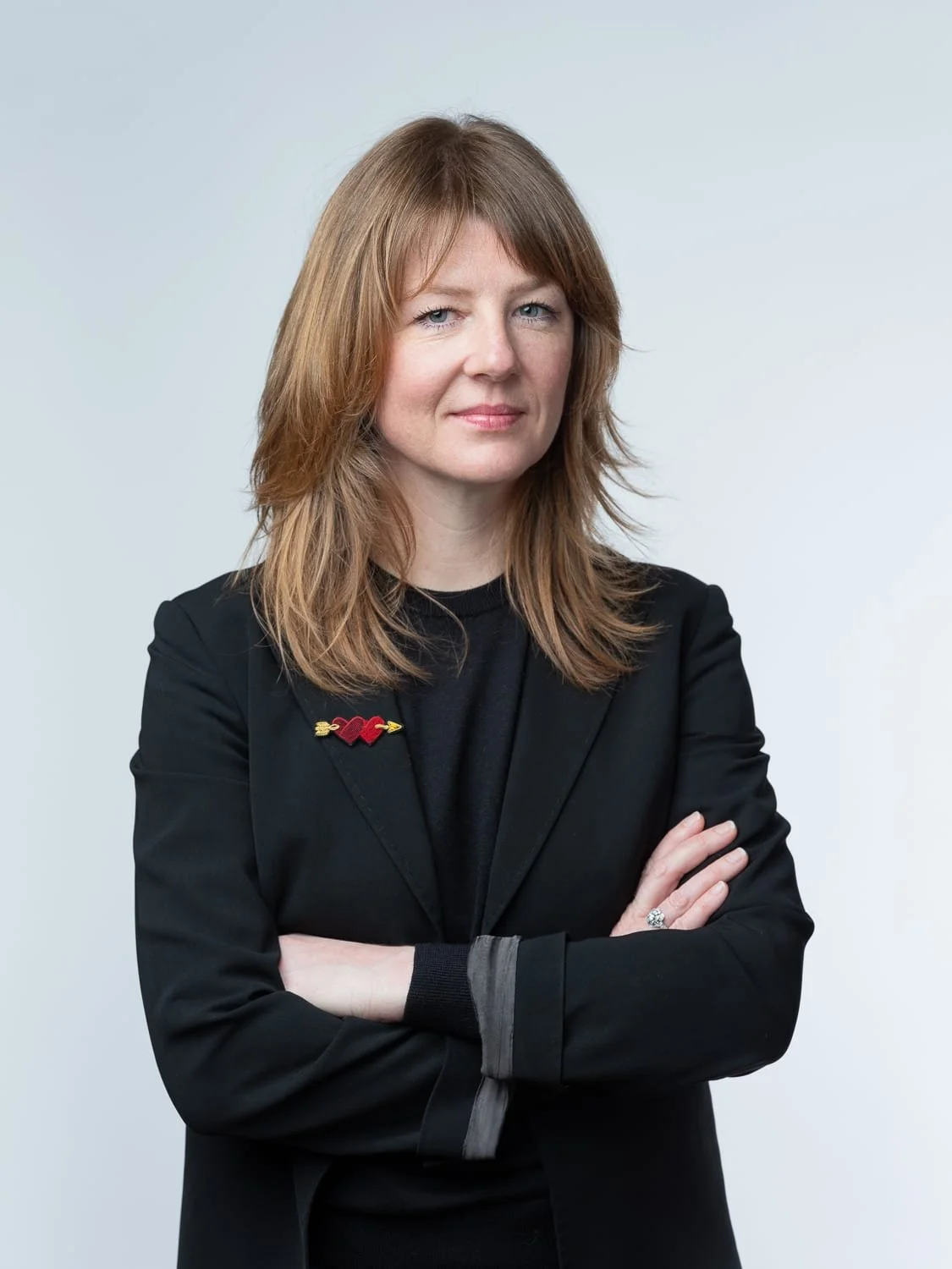 Portrait en studio d'une dirigeante d'entreprise, vêtue d'une veste noire, les bras croisés devant un fond gris clair.