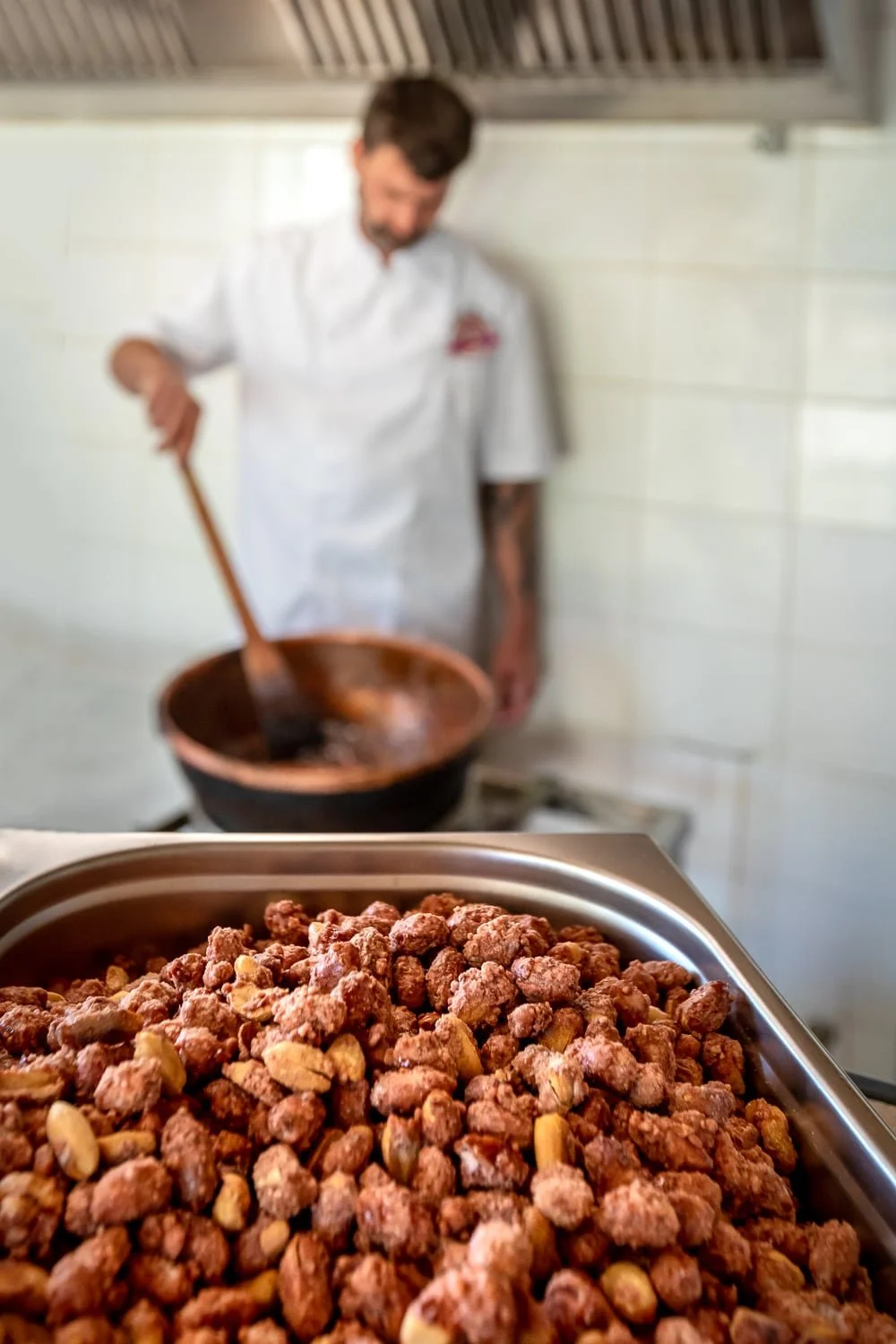 Vue sur un récipient en inox rempli de pralines avec au fond l'artisan qui remue le bassine