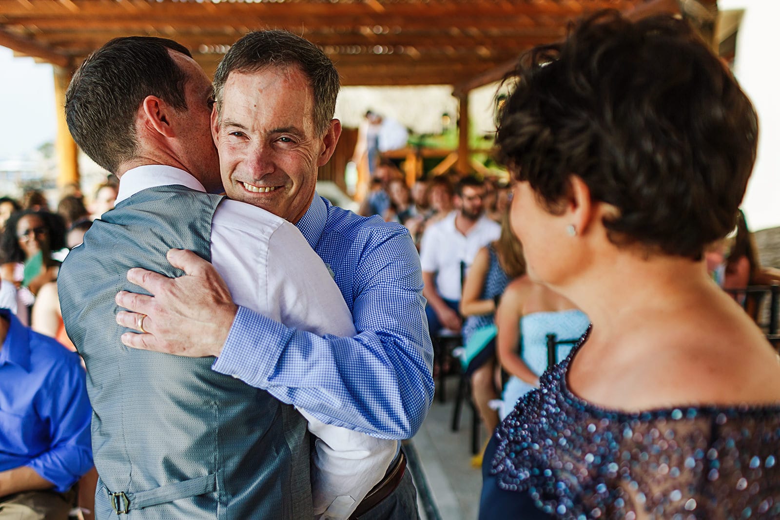 Father hugs groom at the end of the aisle, mom can be seen in foreground