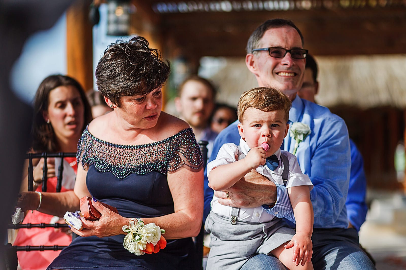 Toddler enjoys a lollipop on father-of-the-groom's lap during wedding ceremony