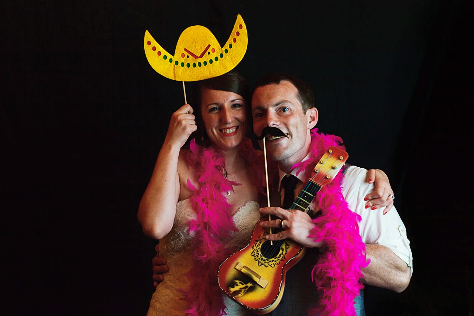 Bride and groom wear a feather boa, fake mustache and sombrero on the photo booth
