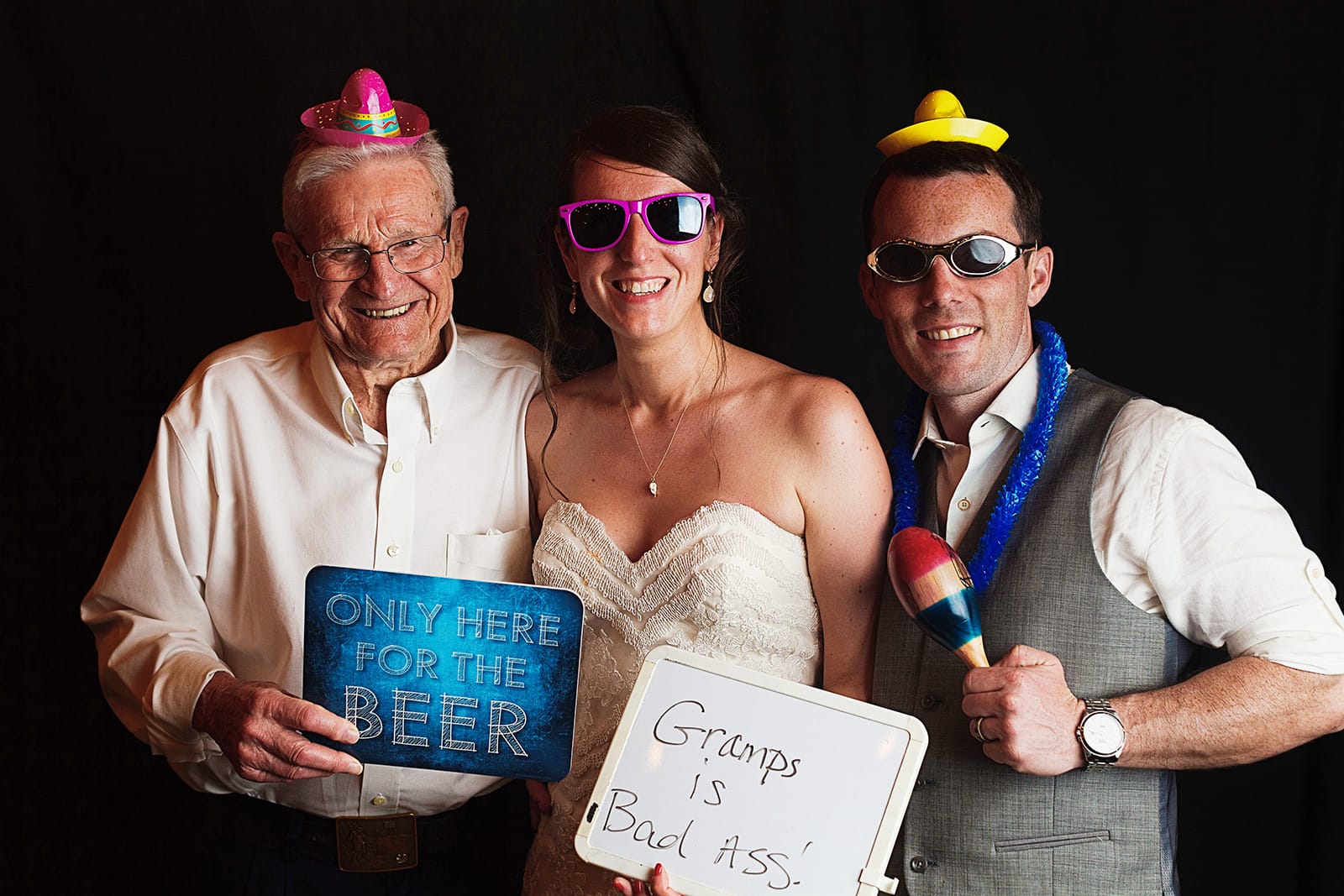 Bride, groom and gramps wear tiny sombreros and hold signs on the photo booth, the bride's one reads "Gramps is Bad Ass"