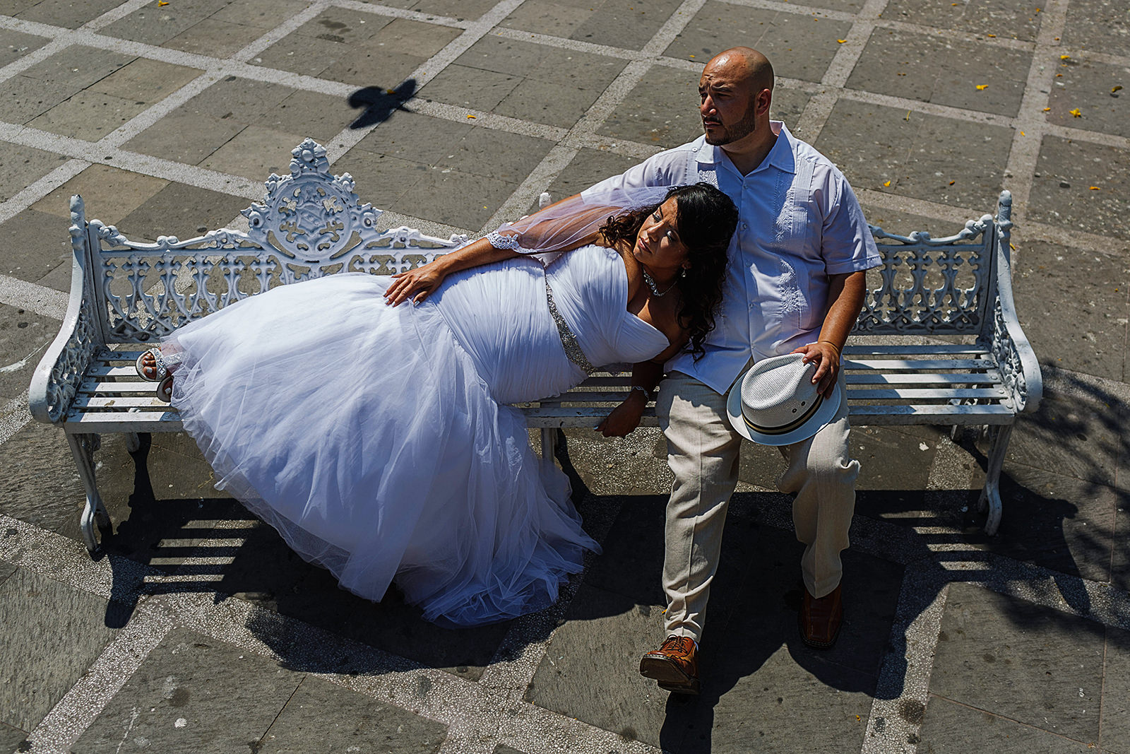 Groom sitting down in a white bench at the main square of Puerto Vallarta, bride is laying on him while a pigeon pass-by