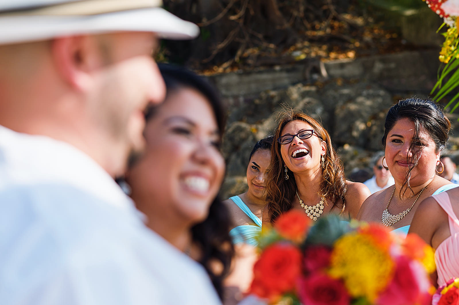 Bridesmaid laughs during beach wedding ceremony, bride and groom can be seen in foreground