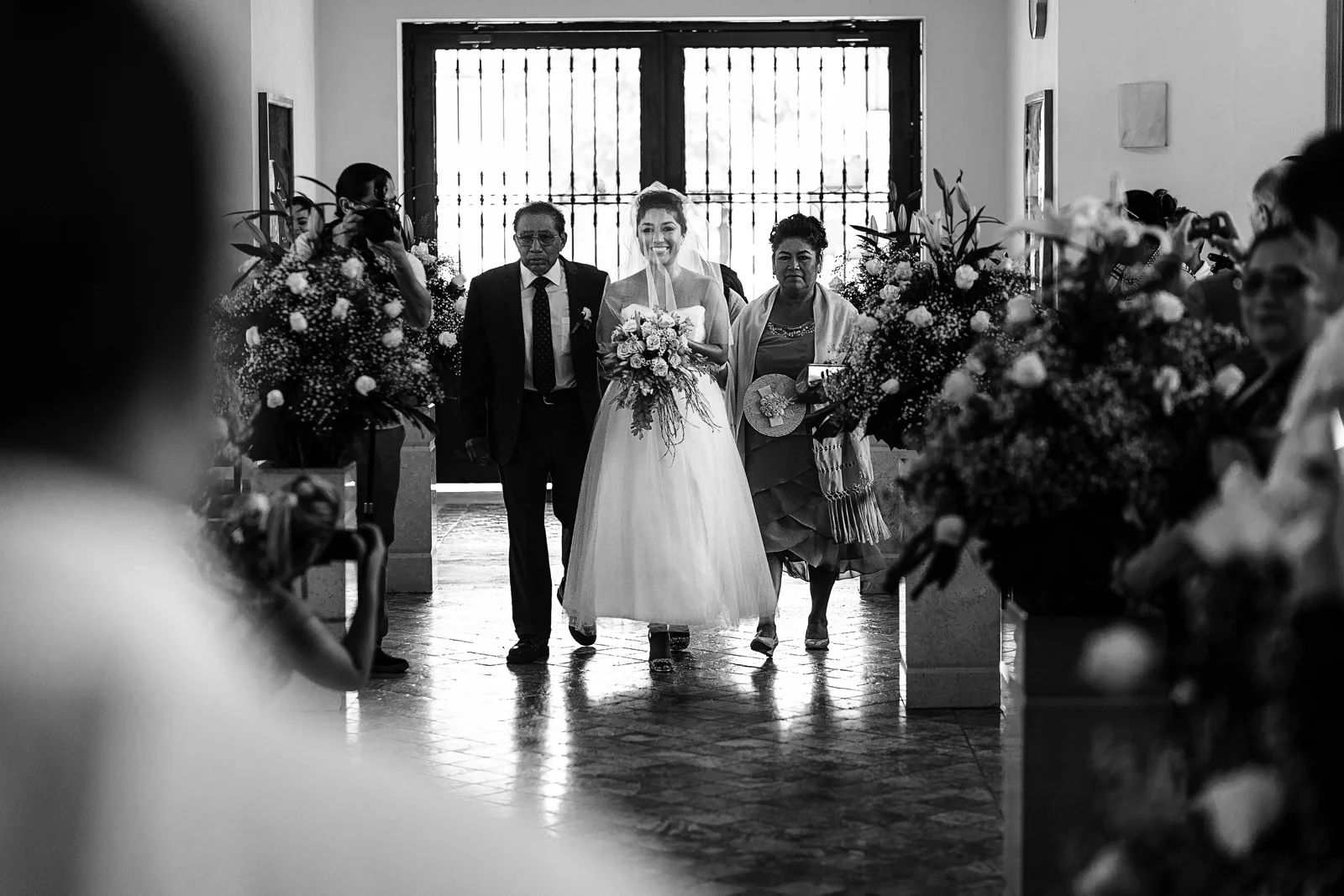 Novia sonriente caminando hacia el altar con sus padres a los lados en el día de su boda