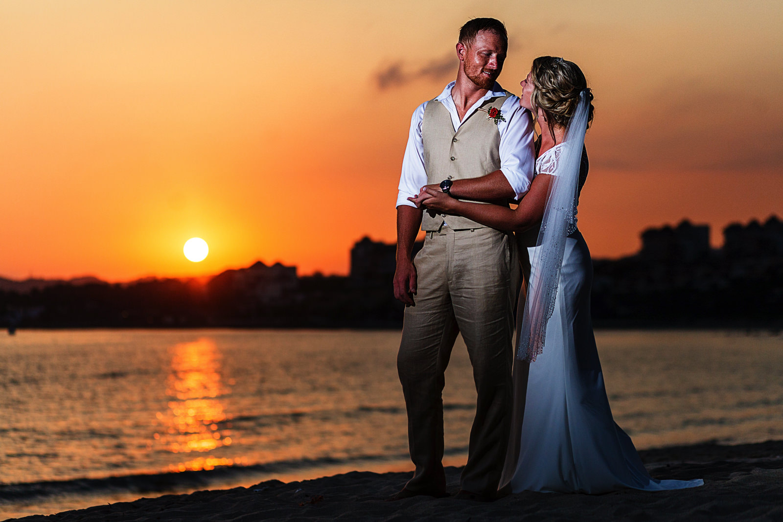 Wedding couple's portrait standing on the beach at Martoca Beach Garden with sunset behind them