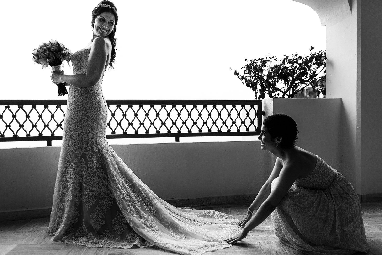 Sister of the bride fixing the train of the wedding dress while the bride is standing holding the flowers outside the bridal suite