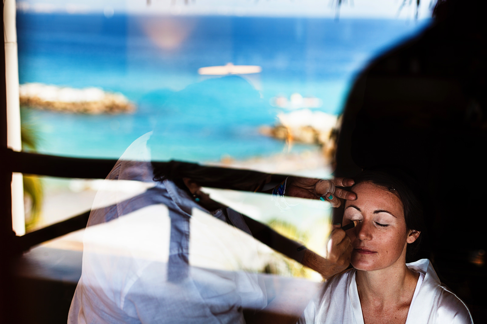 Bride having her make-up in front of a window with the reflection of the ocean