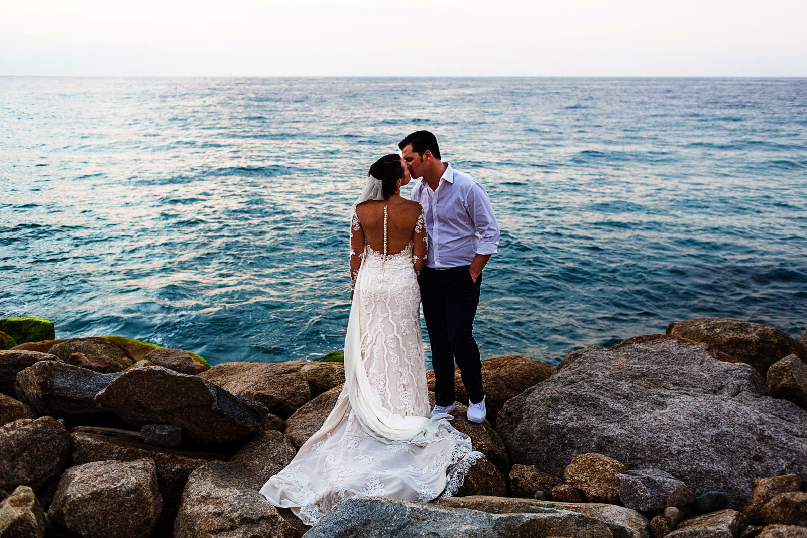 Groom and bride standing on a pile of rocs kissing with the ocean on their background