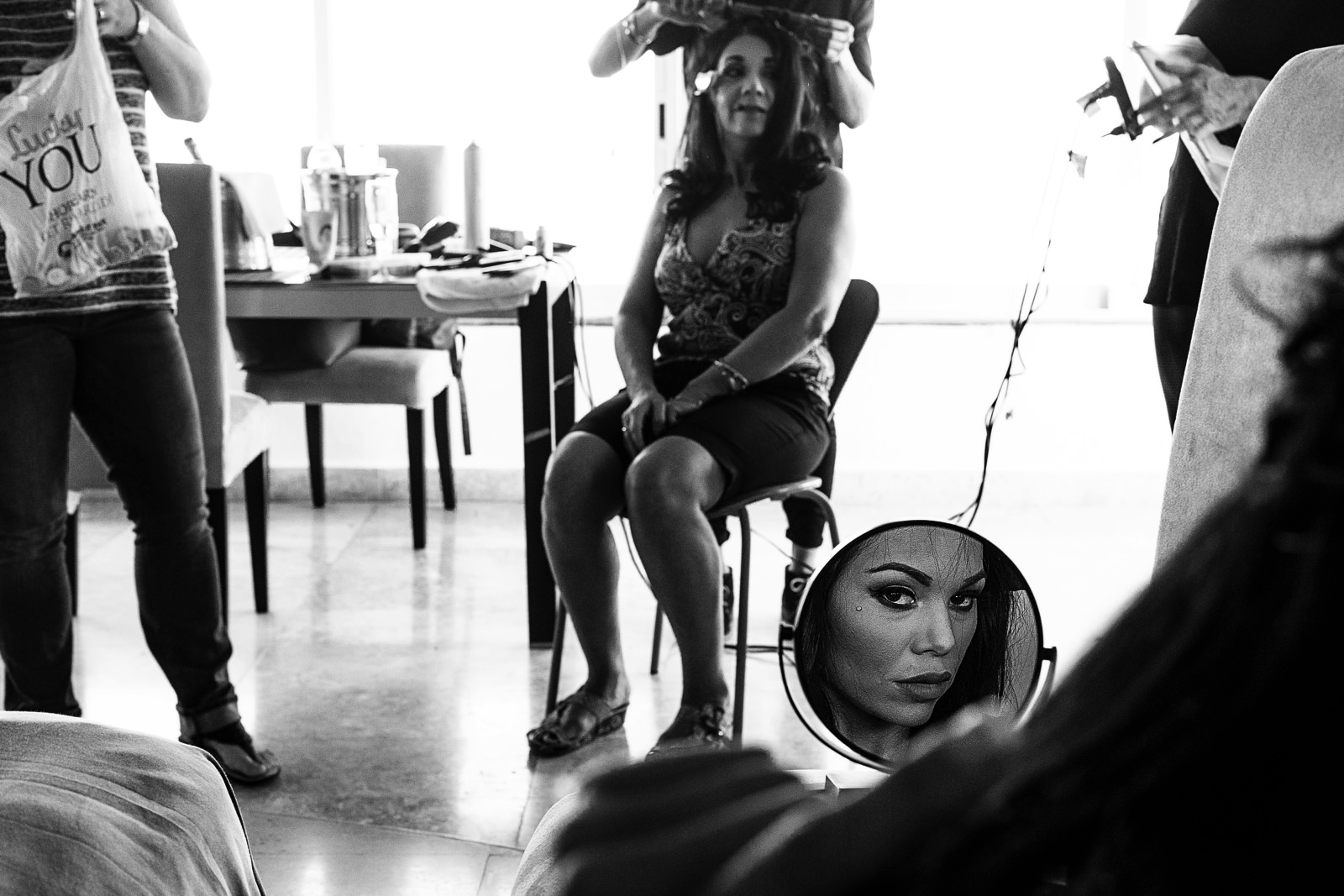 The bride's face is on the mirror and at the background her mother is getting her hair done