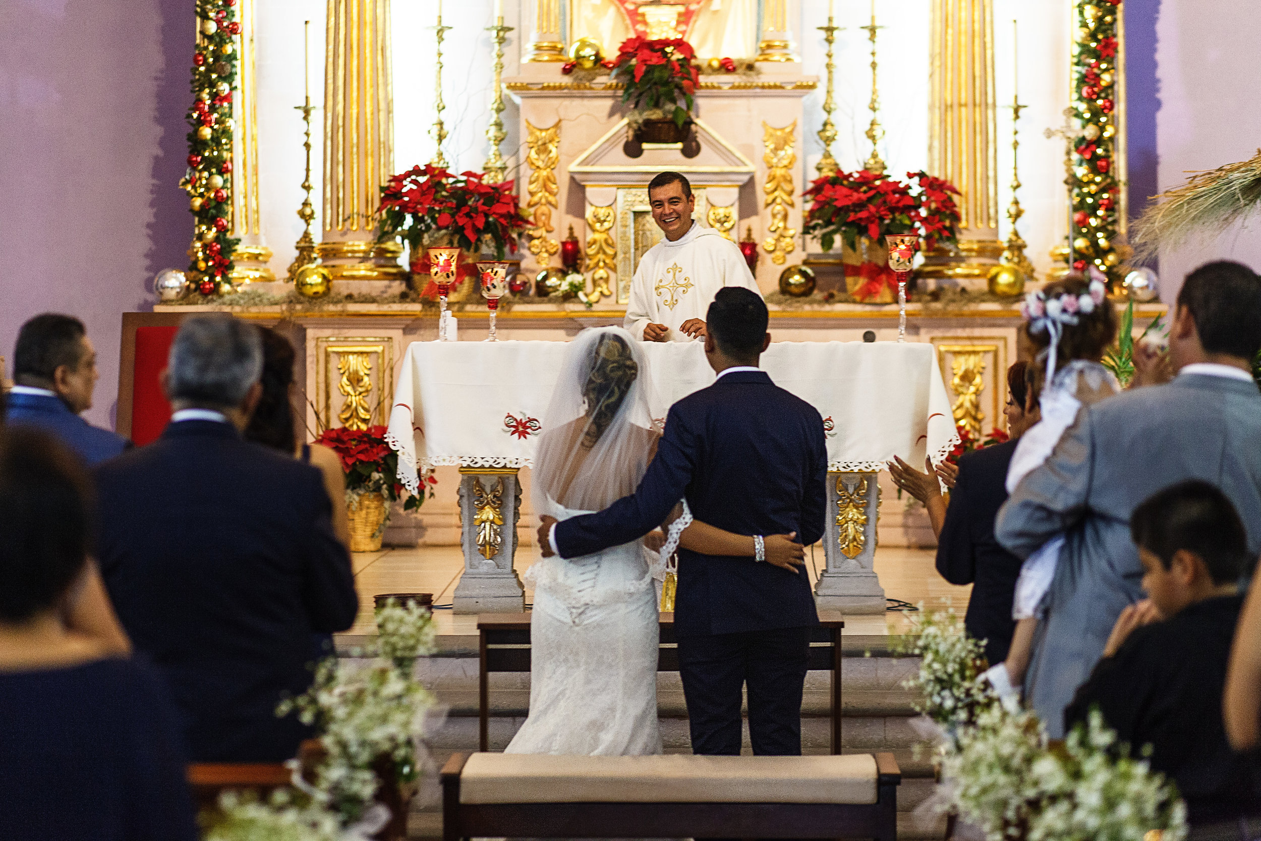 El sacerdote de la iglesia ríe en su sermon frente a la pareja de recién casados que permanecen abrazados frente al altar