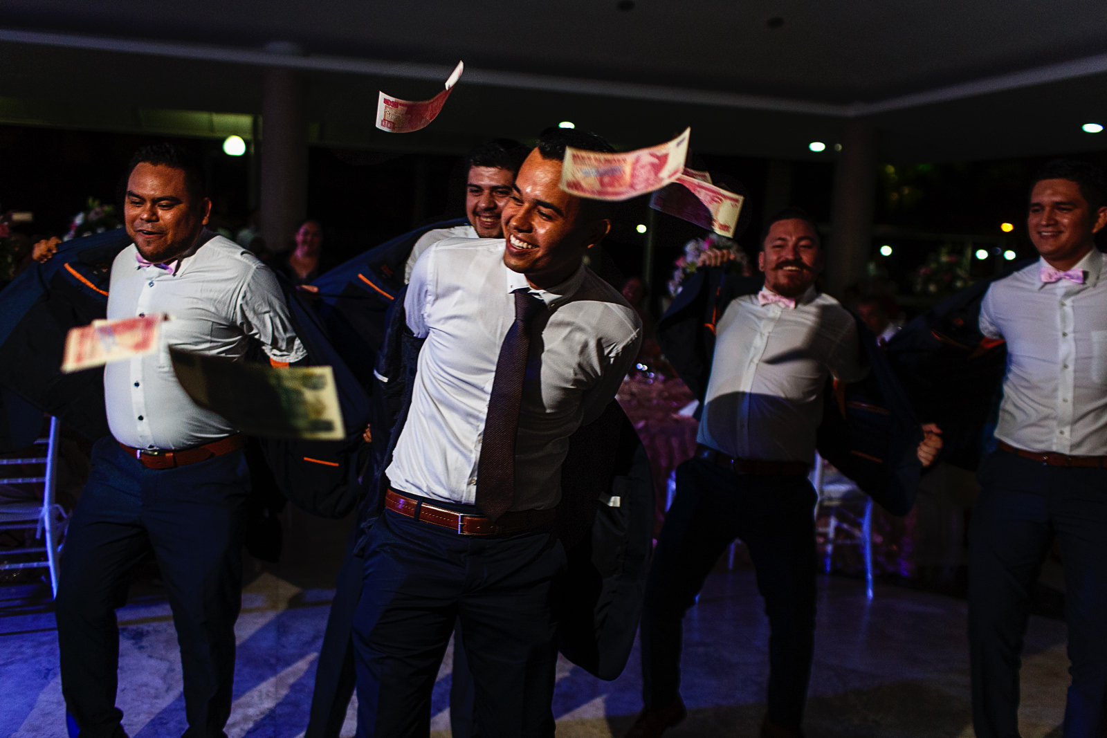 Novio y amigos bailan y se quitan el saco del traje mientras hay billetes volando arrojados por las damas durante la fiesta de la boda