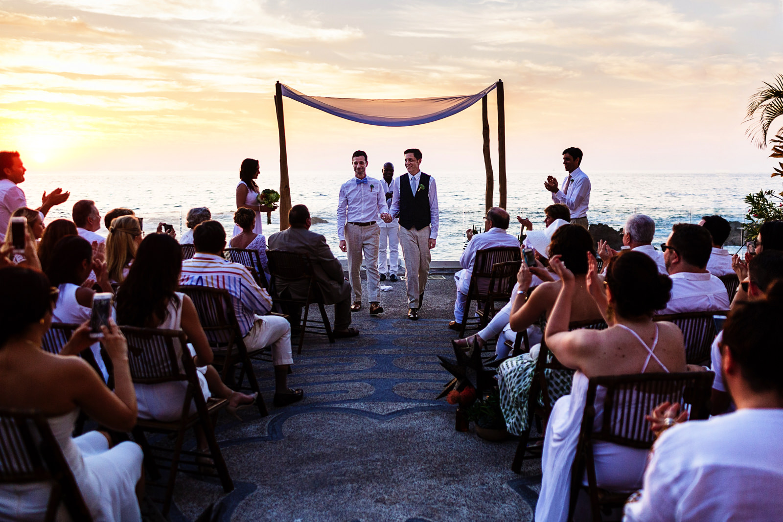 Gay couple exit down the aisle as their ceremony ends with the sunset behind them