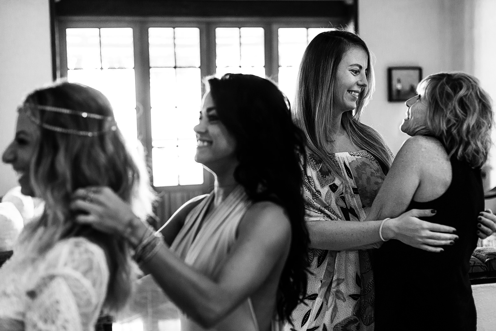 Mother of the bride and a close friend hug behind the bride and sister fixing their hair