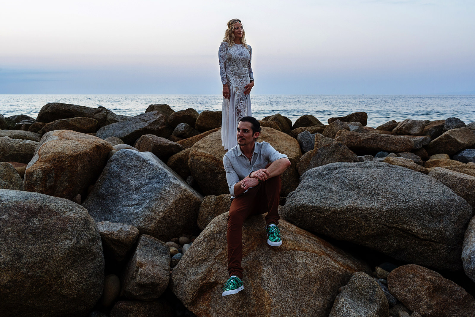 Wedding couple portrait, bride standing in the rocks while the groom is sitting on the rocks with the Mexican pacific ocean in the background