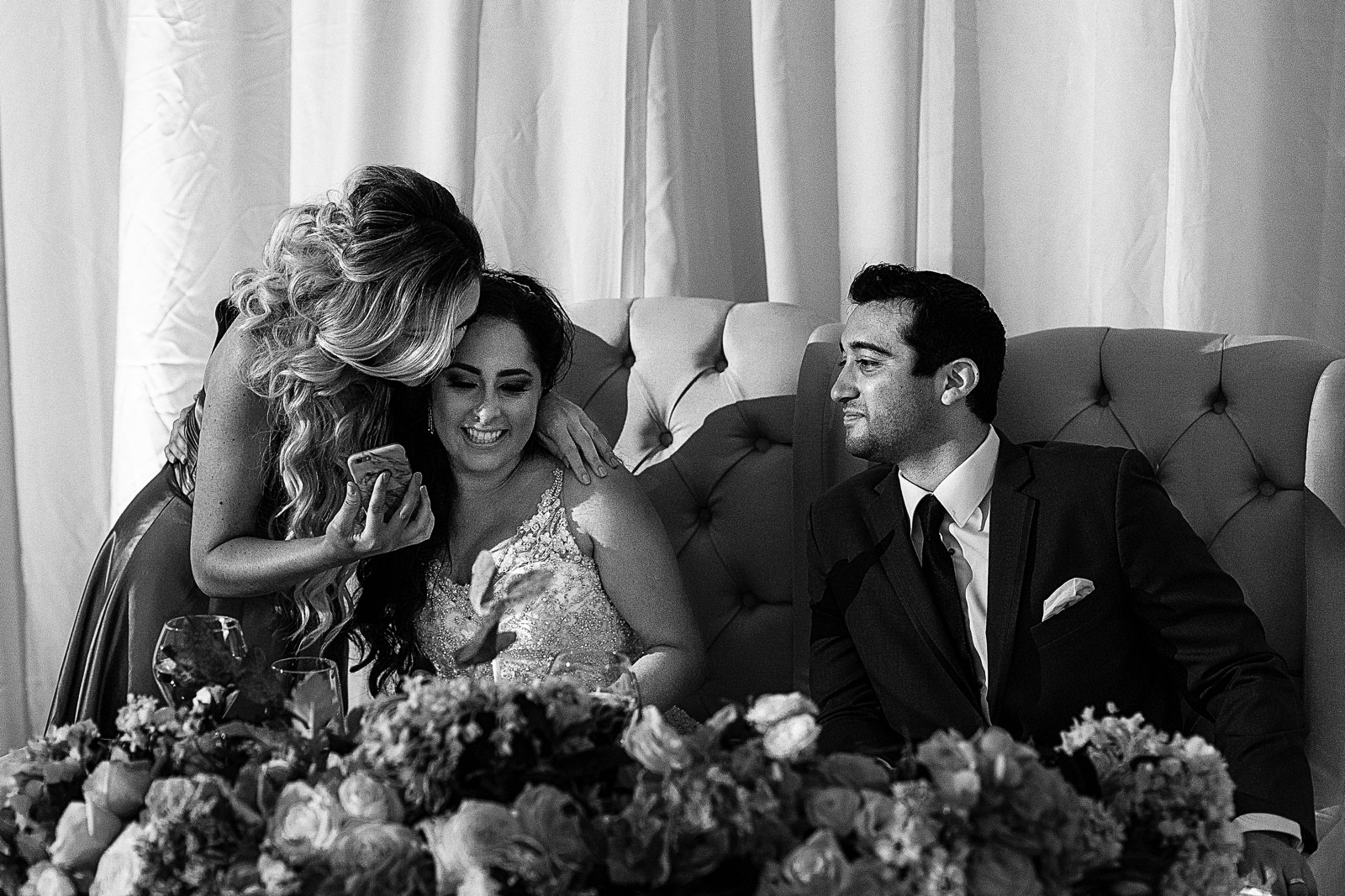 A bridesmaid hugs the bride and shows her a photo on her phone.