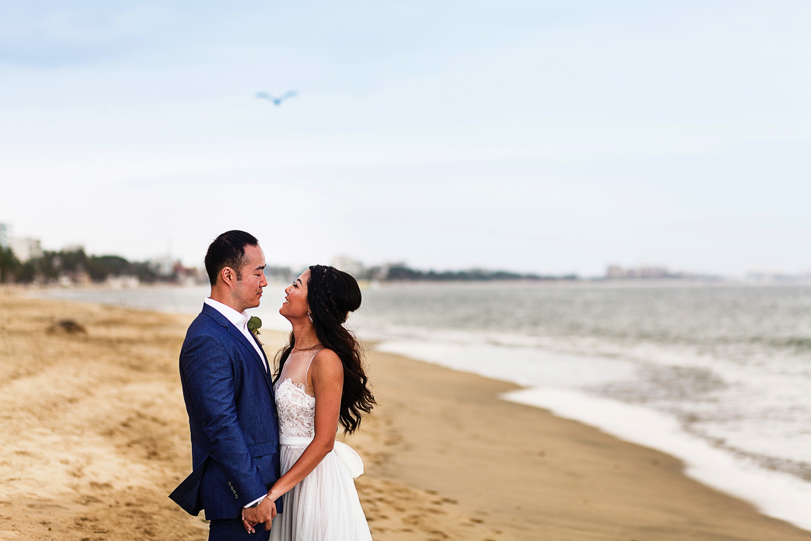 Groom and bride standing on the beach at Bucerias near the ocean with a pelican flying above them.