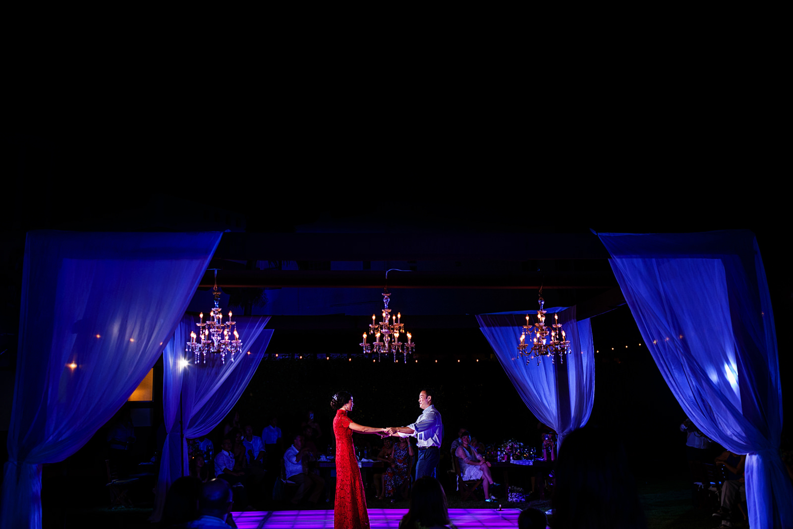 Groom and bride have their first dance as a married couple at the dance floor under a structure with chandeliers.