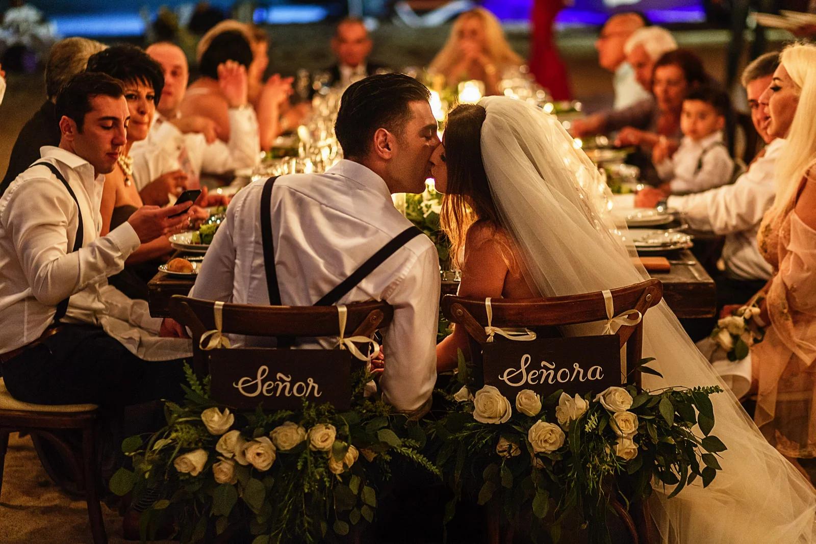 Bride and groom kiss while sitting at their chairs with hanging signs with words "Señor" and "Señora"